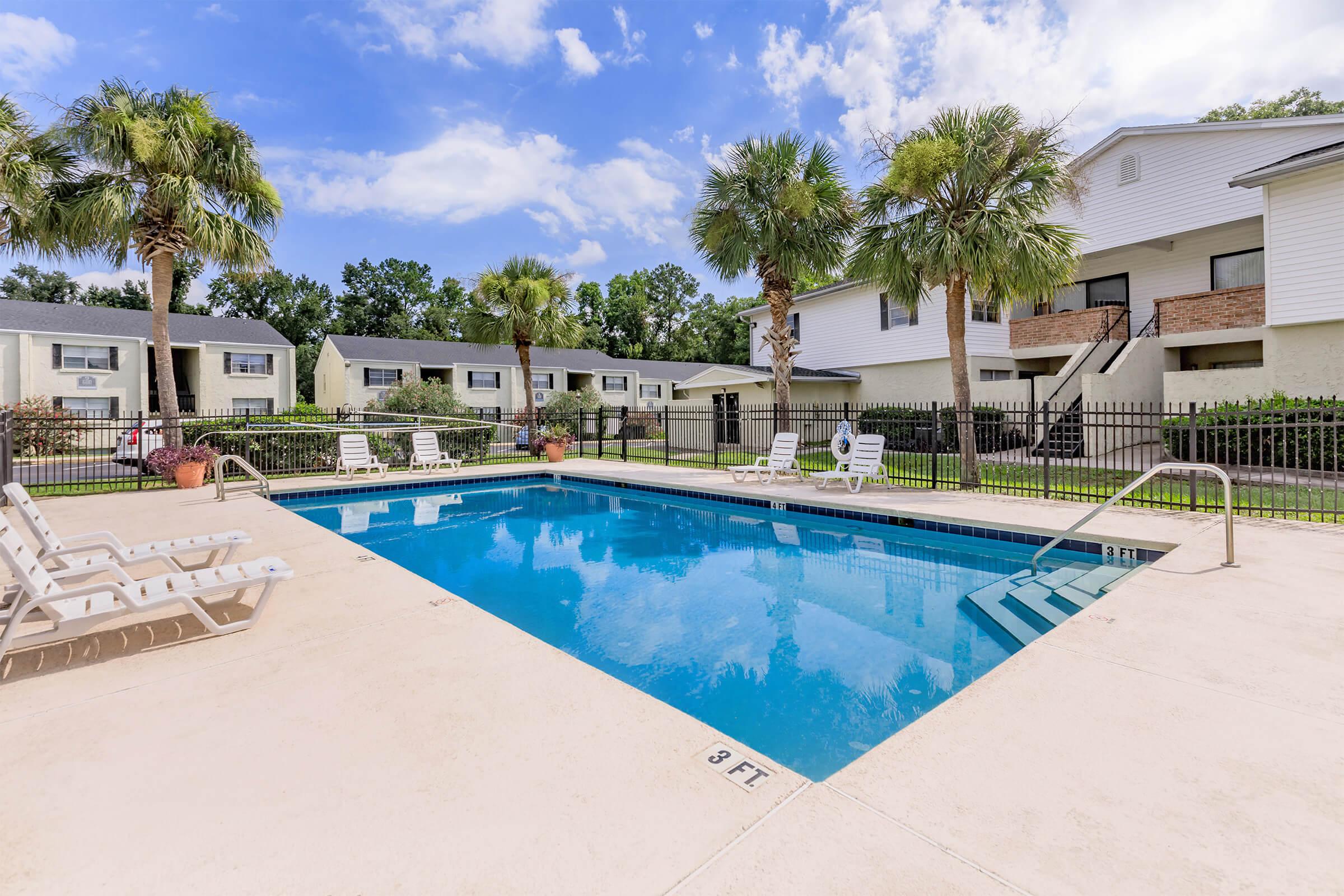 A serene swimming pool surrounded by palm trees, with white lounge chairs placed around it. In the background, there are multiple residential buildings with a clear blue sky overhead and a few fluffy clouds. The pool area is fenced for privacy.