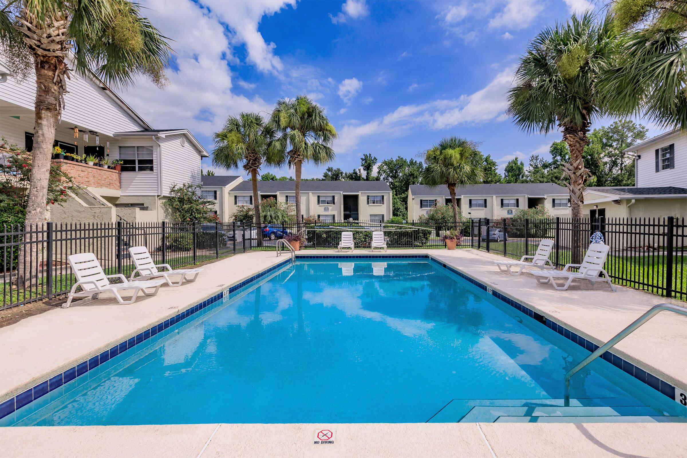 A clear swimming pool surrounded by lounge chairs and palm trees under a blue sky with scattered clouds. In the background, there are white apartment buildings, and a grassy area is visible. A no diving sign is placed near the pool steps.