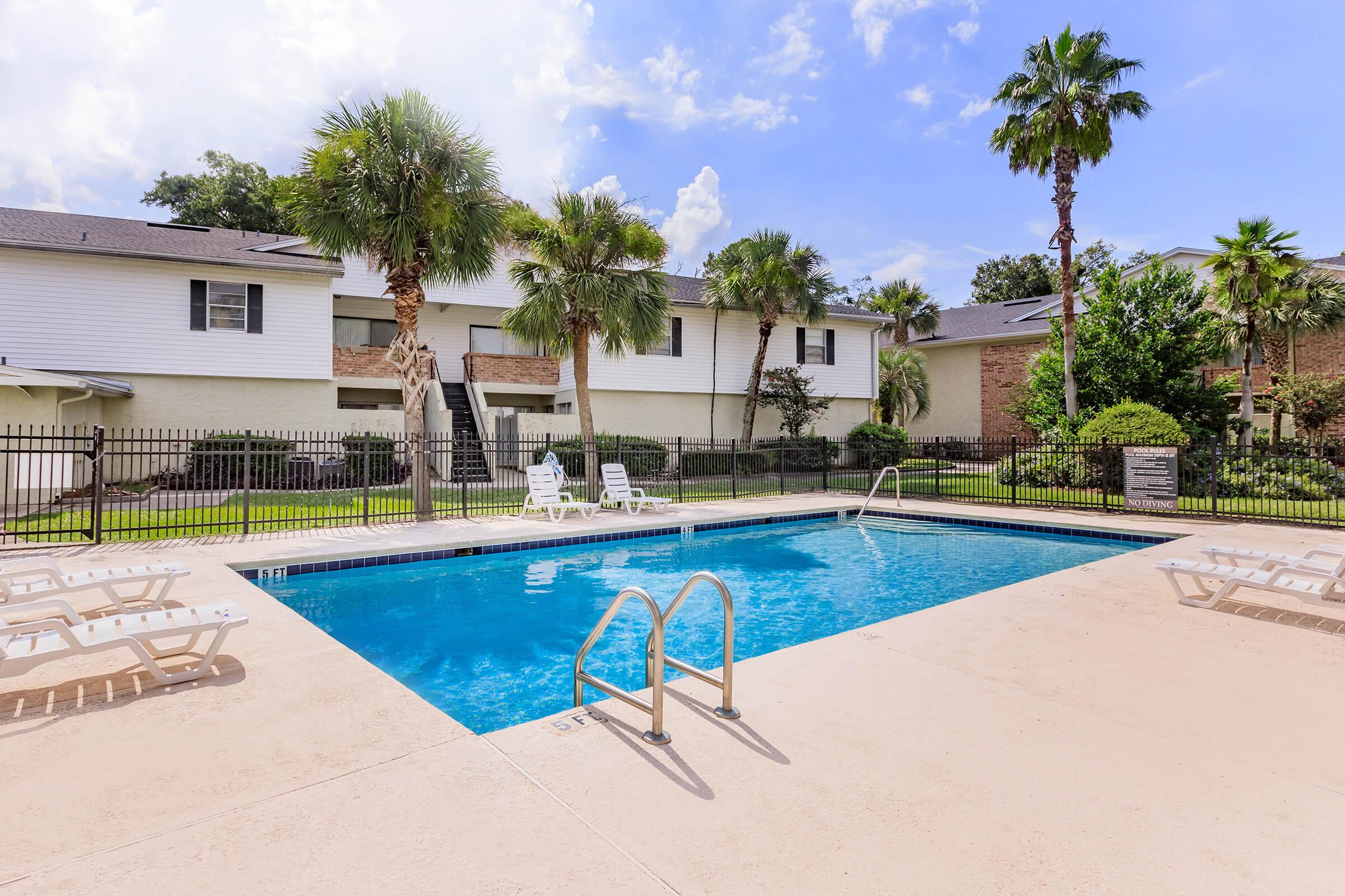 Swimming pool in a landscaped area featuring palm trees and lounge chairs. Surrounding the pool are residential buildings with greenery. The sky is partly cloudy.