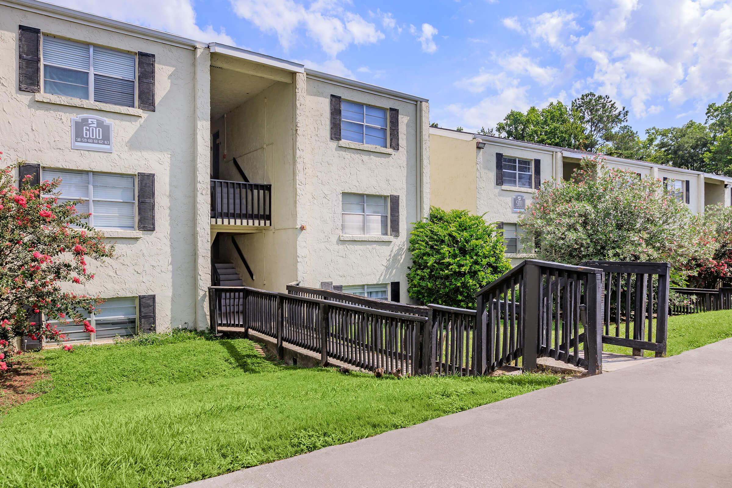 Multistory apartment building with white exterior and black shutters, featuring a wooden walkway leading to a set of stairs. Lush green lawns and landscaped shrubs are visible in front of the building, under a bright blue sky with scattered clouds.