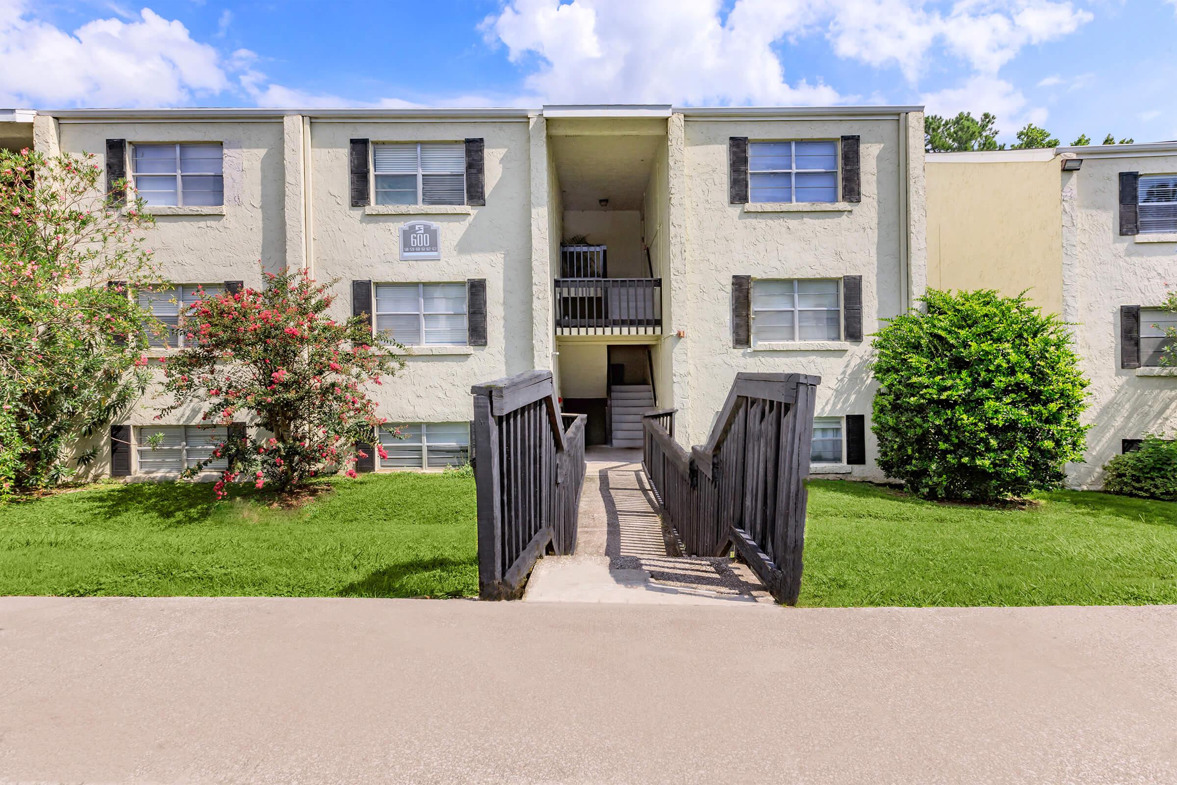 A two-story apartment building with beige exterior walls and black shutters. A sloped wooden ramp leads to the entrance, which has a covered balcony. Lush green grass and shrubs surround the building, and a clear blue sky with some clouds is visible above.