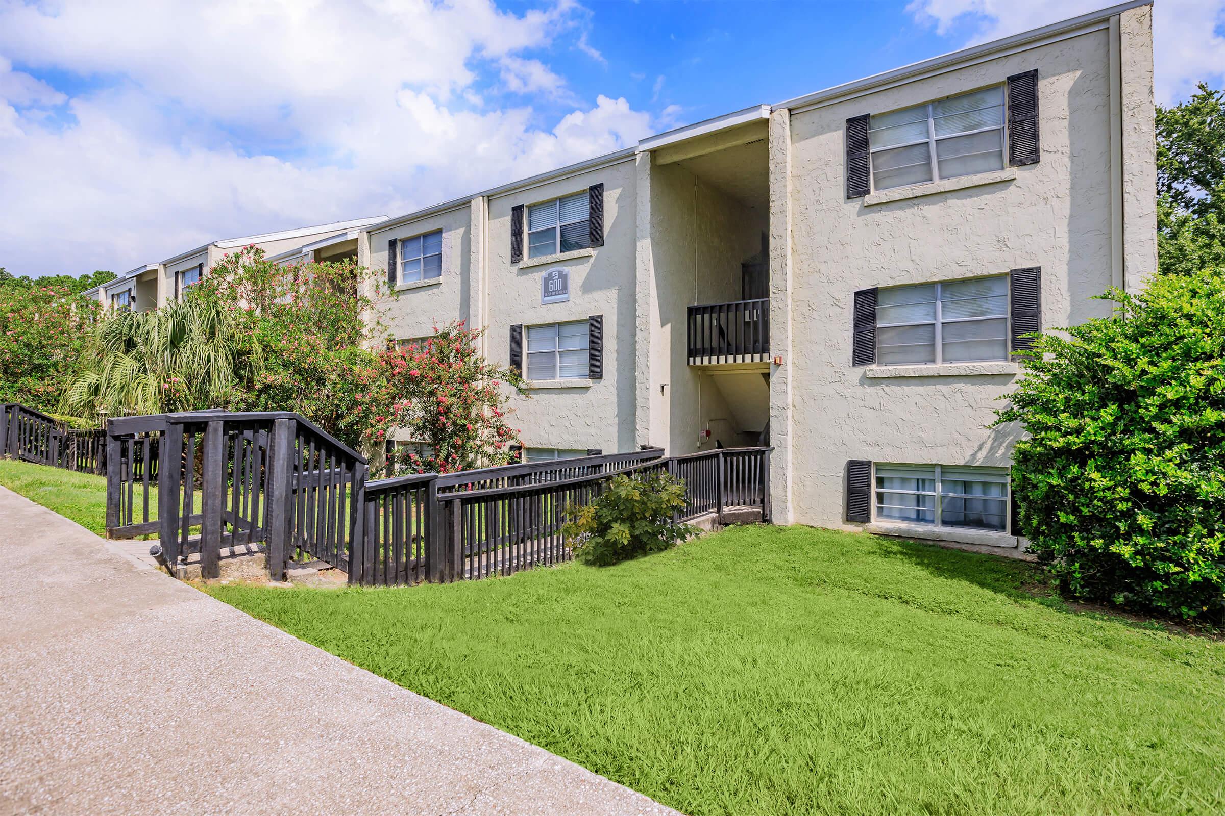 Two-story apartment building with light-colored exterior, black shutters, and a small balcony. The surrounding area features green grass and shrubs, with a pathway leading to the entrance. The sky is partly cloudy, suggesting a bright and sunny day.