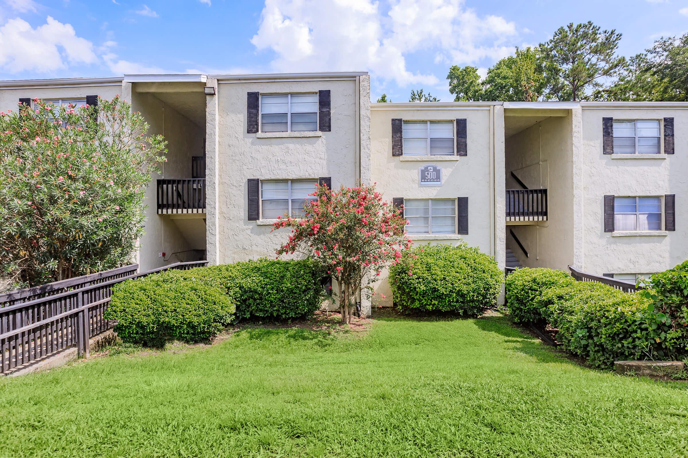 Two-story apartment building with a light-colored exterior, featuring multiple windows with black shutters. In front, there's a well-maintained green lawn and neatly trimmed hedges. A small flowering tree adds a pop of color, and the sky is partly cloudy.
