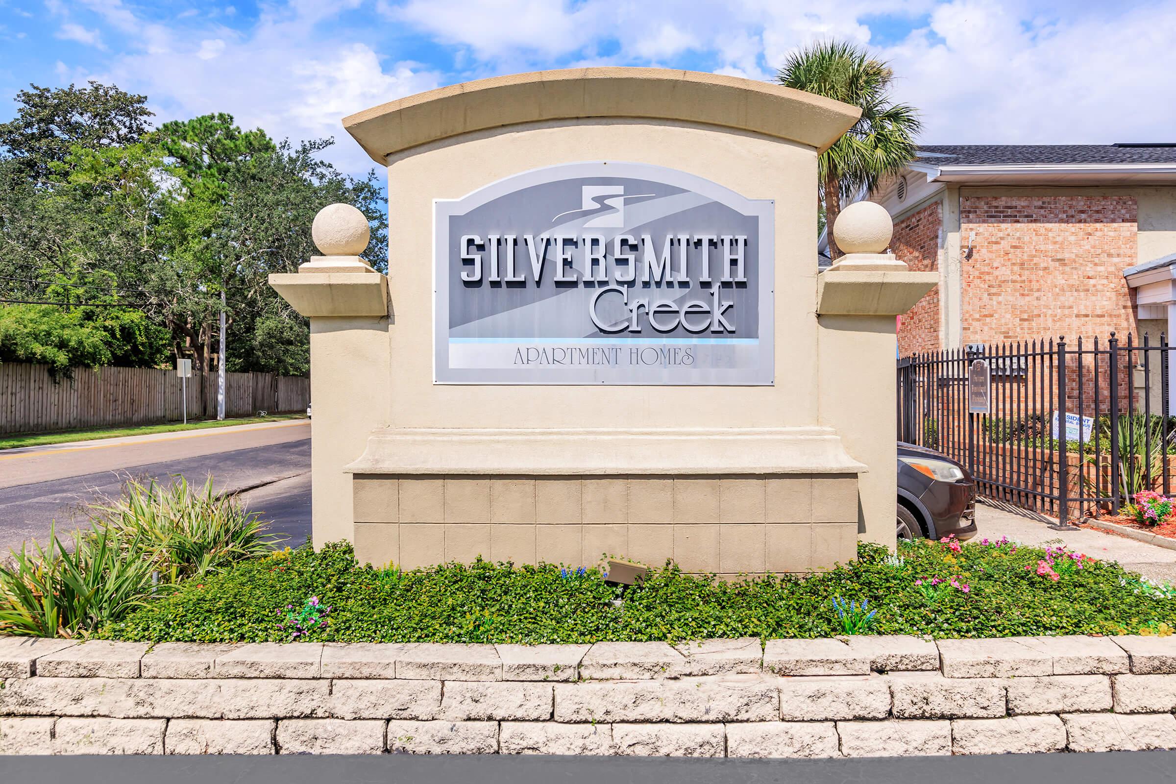 Sign for Silversmith Creek Apartment Homes, featuring the name prominently displayed. The sign is framed by landscaping with greenery and flowers, set against a clear blue sky. The entrance appears well-maintained, contributing to a welcoming atmosphere for residents and visitors.
