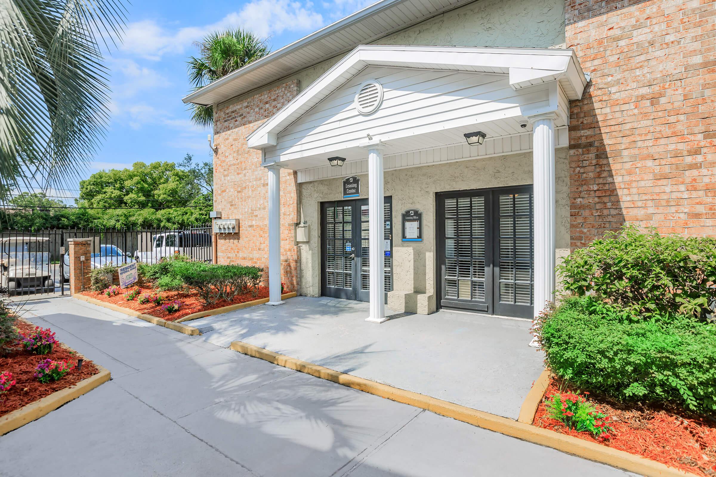 Exterior view of a building entrance featuring a covered porch with white columns. The entrance has double doors, with planters filled with colorful flowers on either side. The pathway is lined with red mulch and greenery, and there are palm trees and a clear blue sky in the background.