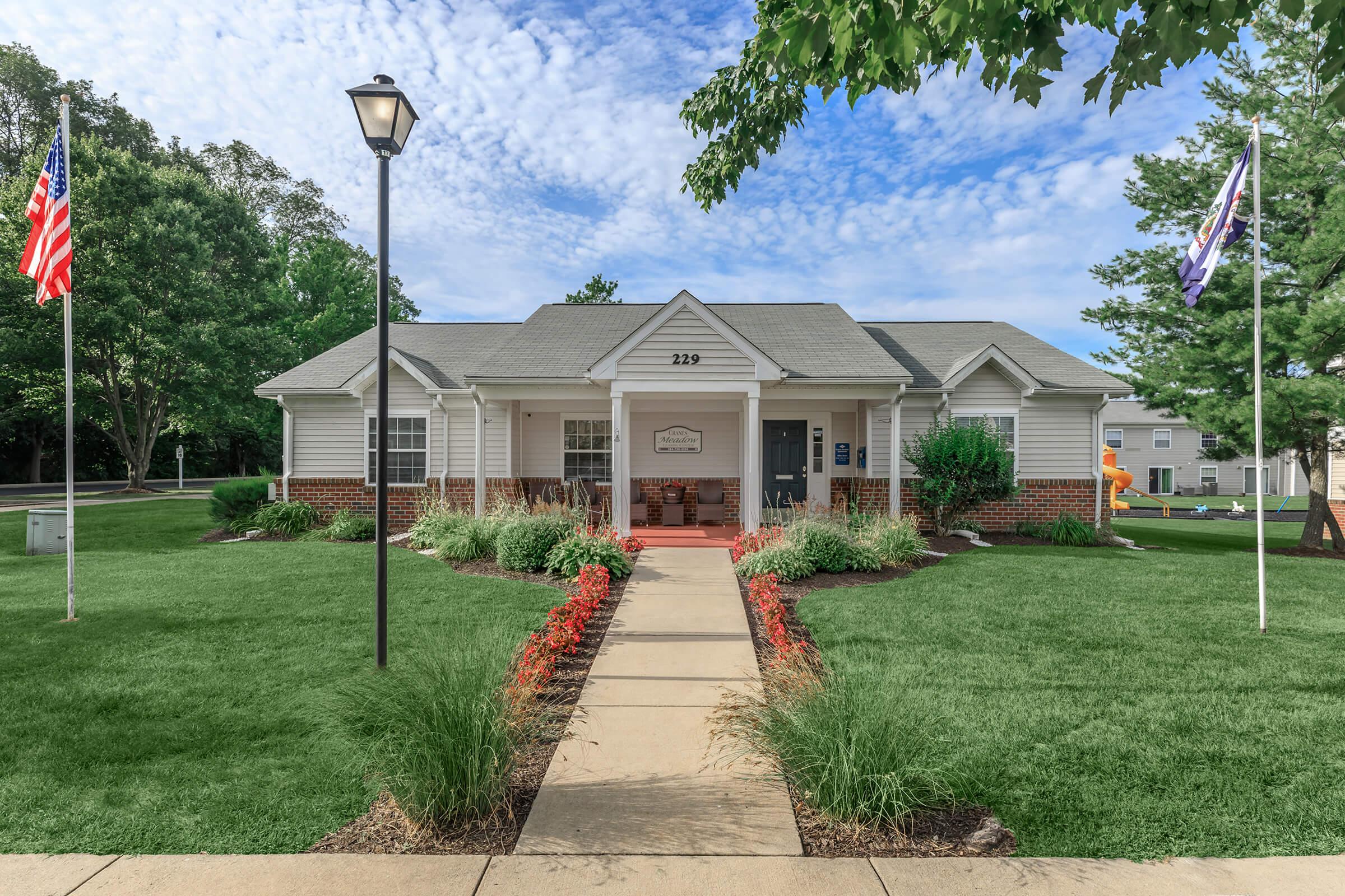 A welcoming single-story building with a light-colored facade and a sloped roof. The entrance features a pathway bordered by colorful flower beds. Flags are displayed on either side of the entrance, and trees provide greenery in the background under a partly cloudy sky.
