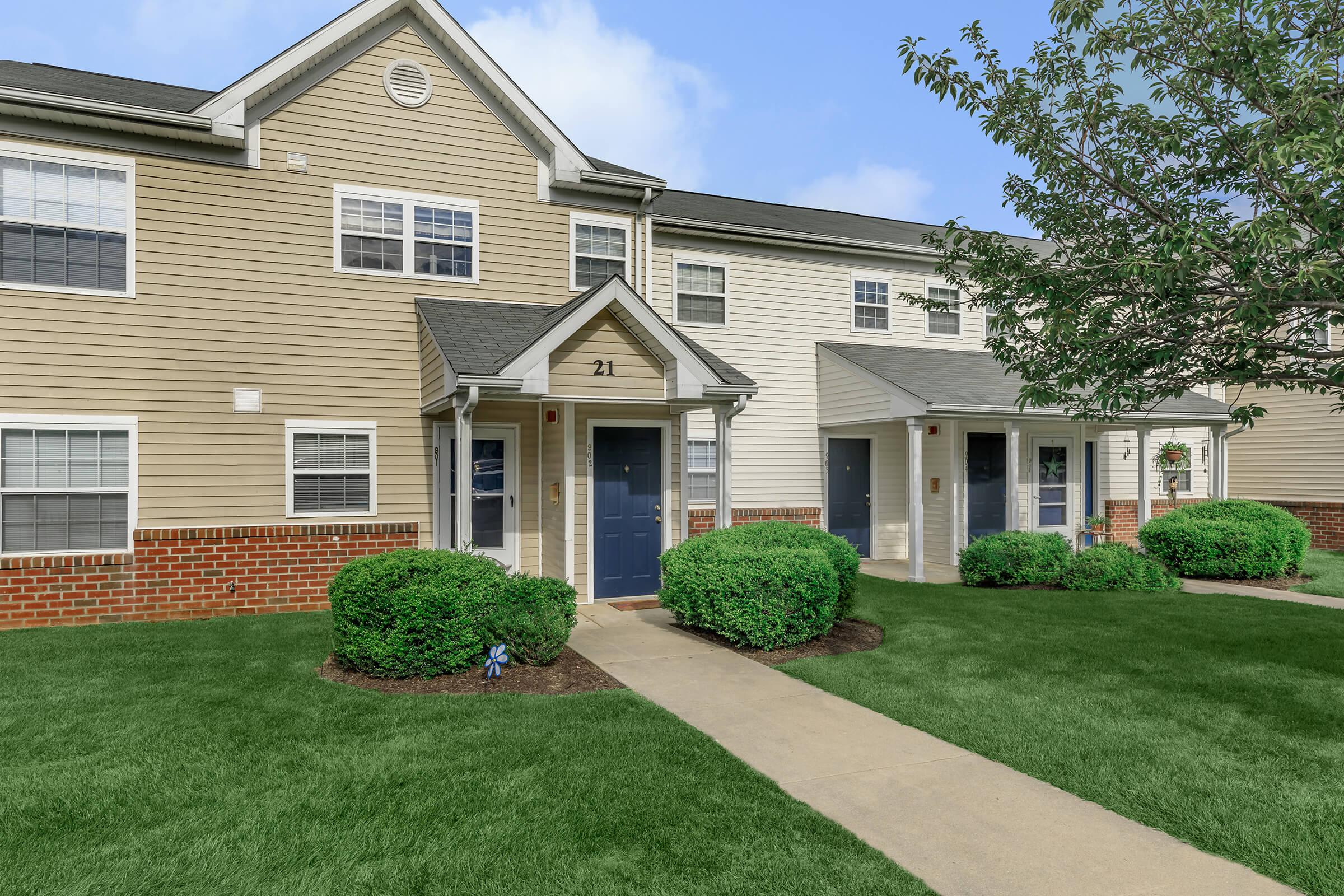 A suburban apartment complex featuring two buildings with a mix of beige and white siding. The entrance doors are painted blue, flanked by neatly trimmed green shrubs and grass. A concrete pathway leads to the doors, under a clear blue sky.