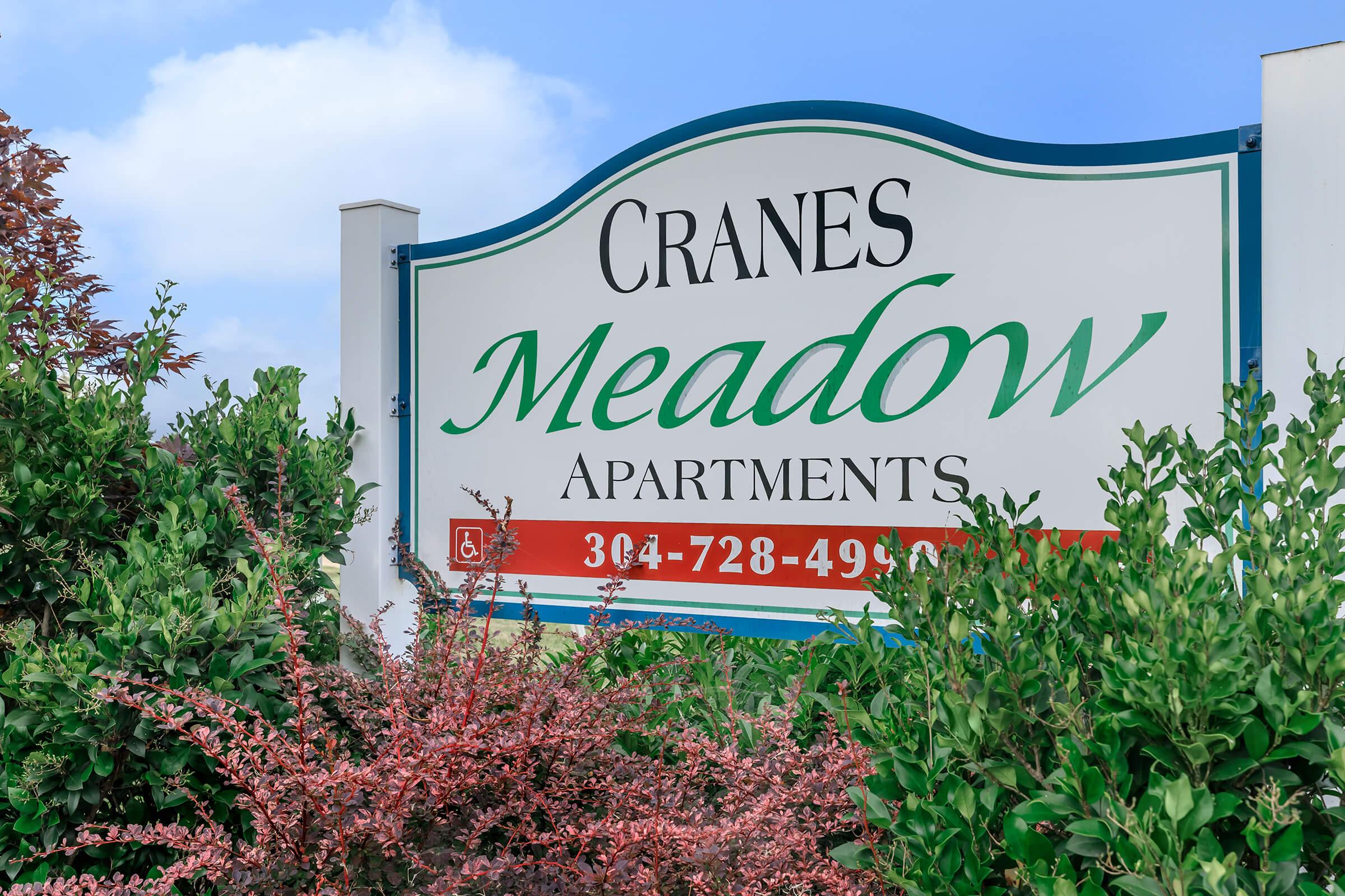 Sign for Cranes Meadow Apartments, featuring green and red text against a white background. Surrounded by lush greenery and colorful bushes, with a blue sky in the background. The sign includes a phone number: 364-728-4999.