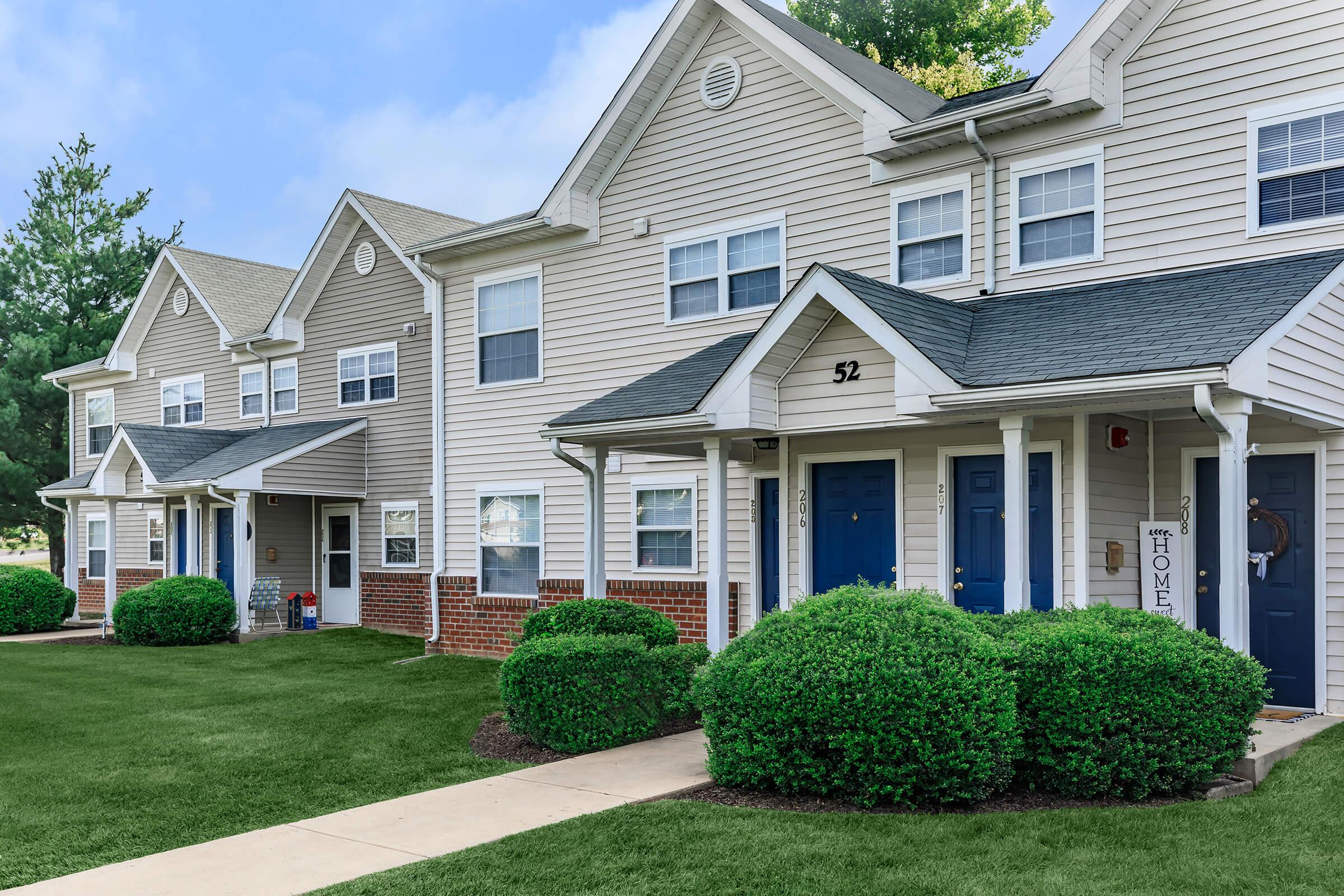A row of two-story townhouses with beige siding and green lawns. Each unit features a blue front door and white windows. The yard is neatly trimmed, and landscaping includes symmetrical bushes. The overall scene reflects a welcoming residential neighborhood atmosphere.