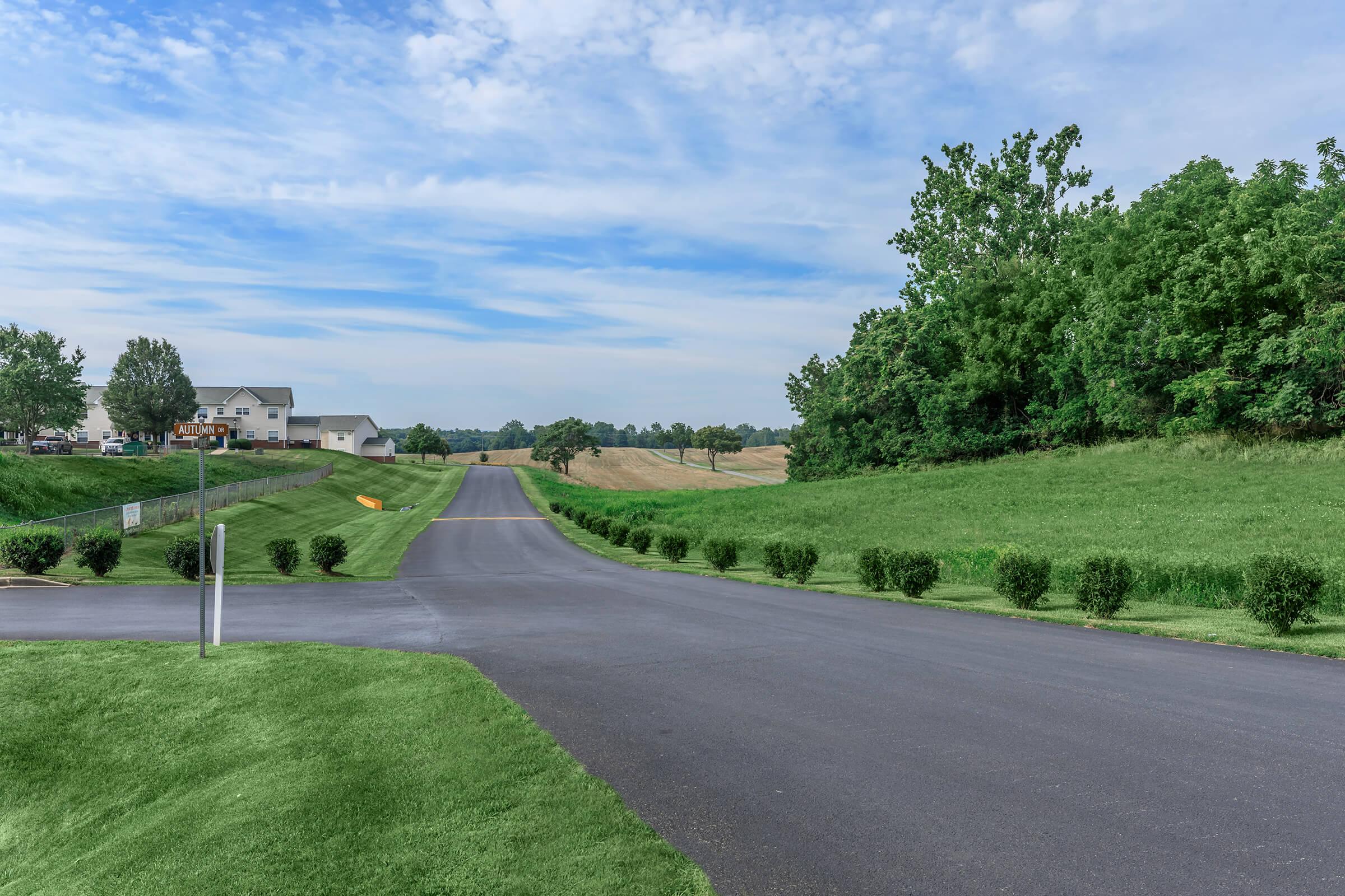 A peaceful rural scene featuring a wide, paved road diverging at a T-intersection. Lush green grass and hedges line the sides of the road, while distant hills are visible under a partly cloudy sky. Nearby, modern houses are set against the backdrop of open fields.