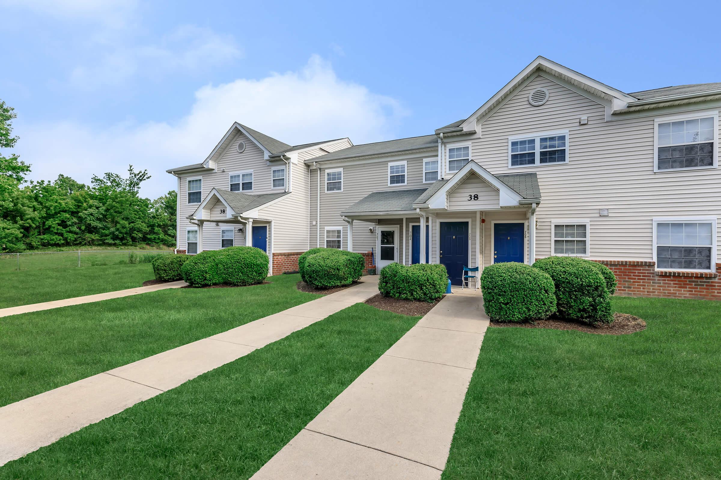 Row of two-story apartment buildings with blue front doors and manicured lawns. Pathways lead to the entrances, and neatly trimmed bushes are visible in front of the units. The sky is clear and blue, creating a pleasant suburban atmosphere.