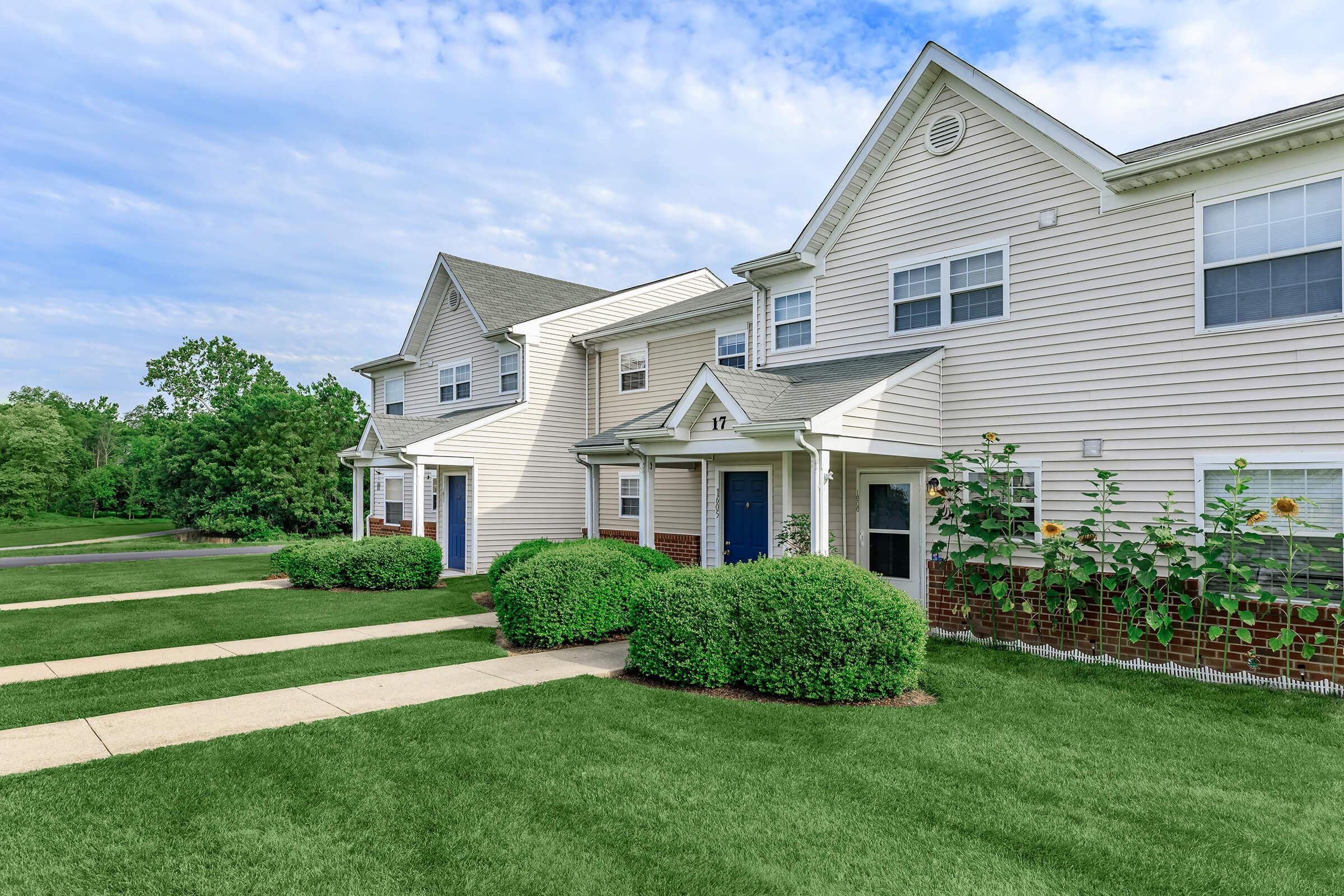 A row of modern townhouse apartments with light-colored siding and contrasting blue doors, set against a lush green lawn. Flowering plants and neatly trimmed bushes line the walkways, while a clear blue sky with scattered clouds provides a picturesque backdrop.
