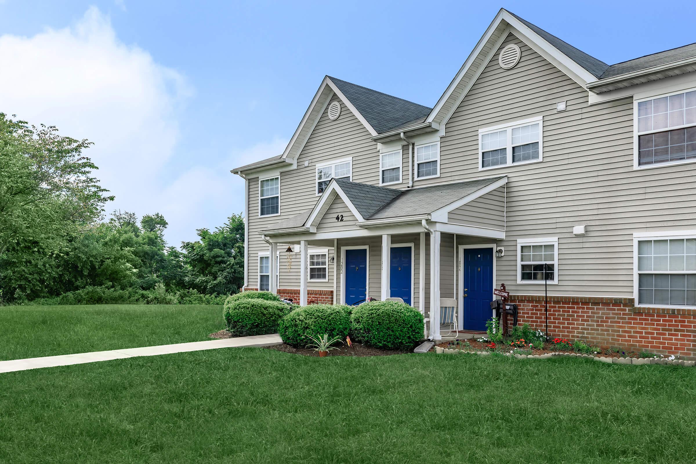 A view of a two-story townhouse with gray siding and blue doors. The front yard features neatly trimmed bushes and a lawn, with a paved walkway leading to the entrance. Behind the townhouse, there are trees and greenery, under a partially cloudy sky.