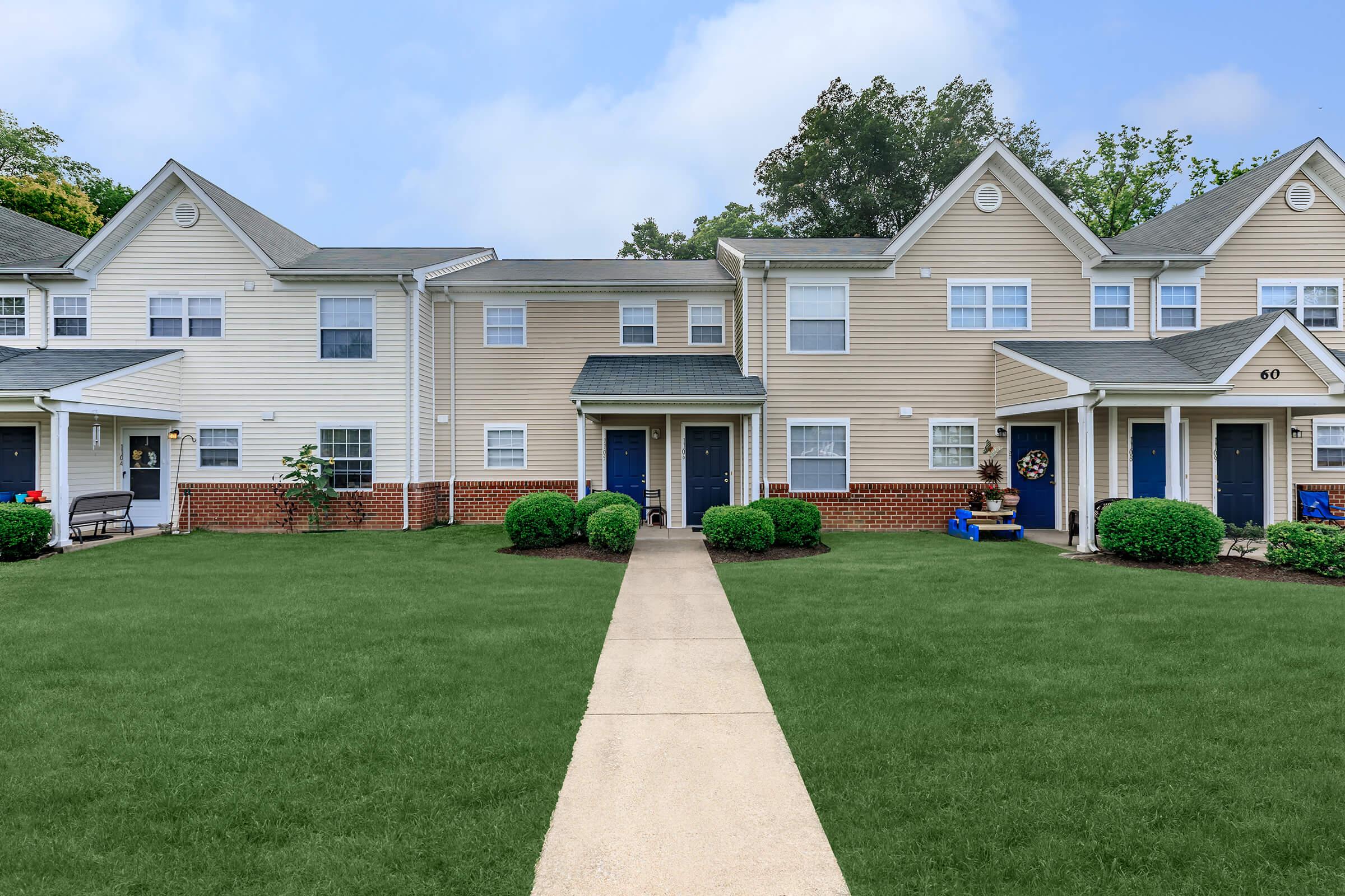 A row of two-story apartment buildings with light-colored siding and blue doors. A neatly maintained lawn with small bushes lines a paved walkway leading to the entrances. The buildings are surrounded by trees and greenery under a cloudy sky.
