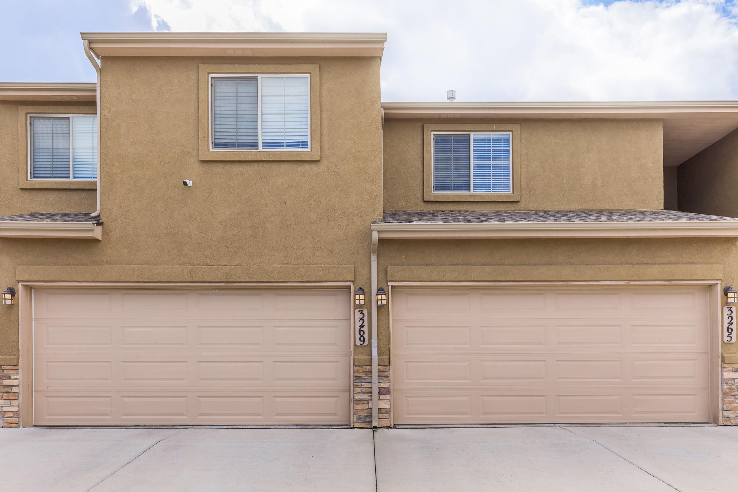 A two-story townhouse or duplex with beige stucco exterior. Each unit has a large garage door with a light fixture beside it. Above the garage, there is a window with horizontal blinds. The sky is partly cloudy, adding a bright atmosphere to the scene.