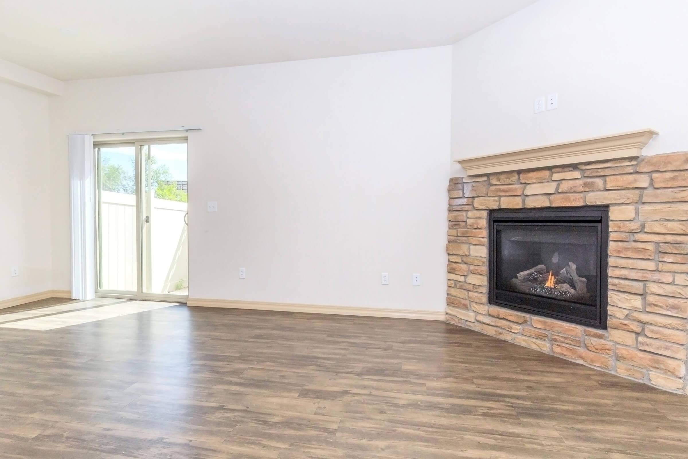 A spacious living room with a stone fireplace on one wall. Large sliding glass doors lead to a balcony, allowing natural light to fill the space. The walls are painted in a light color, and the floor is made of dark wood laminate, creating a warm and inviting atmosphere.