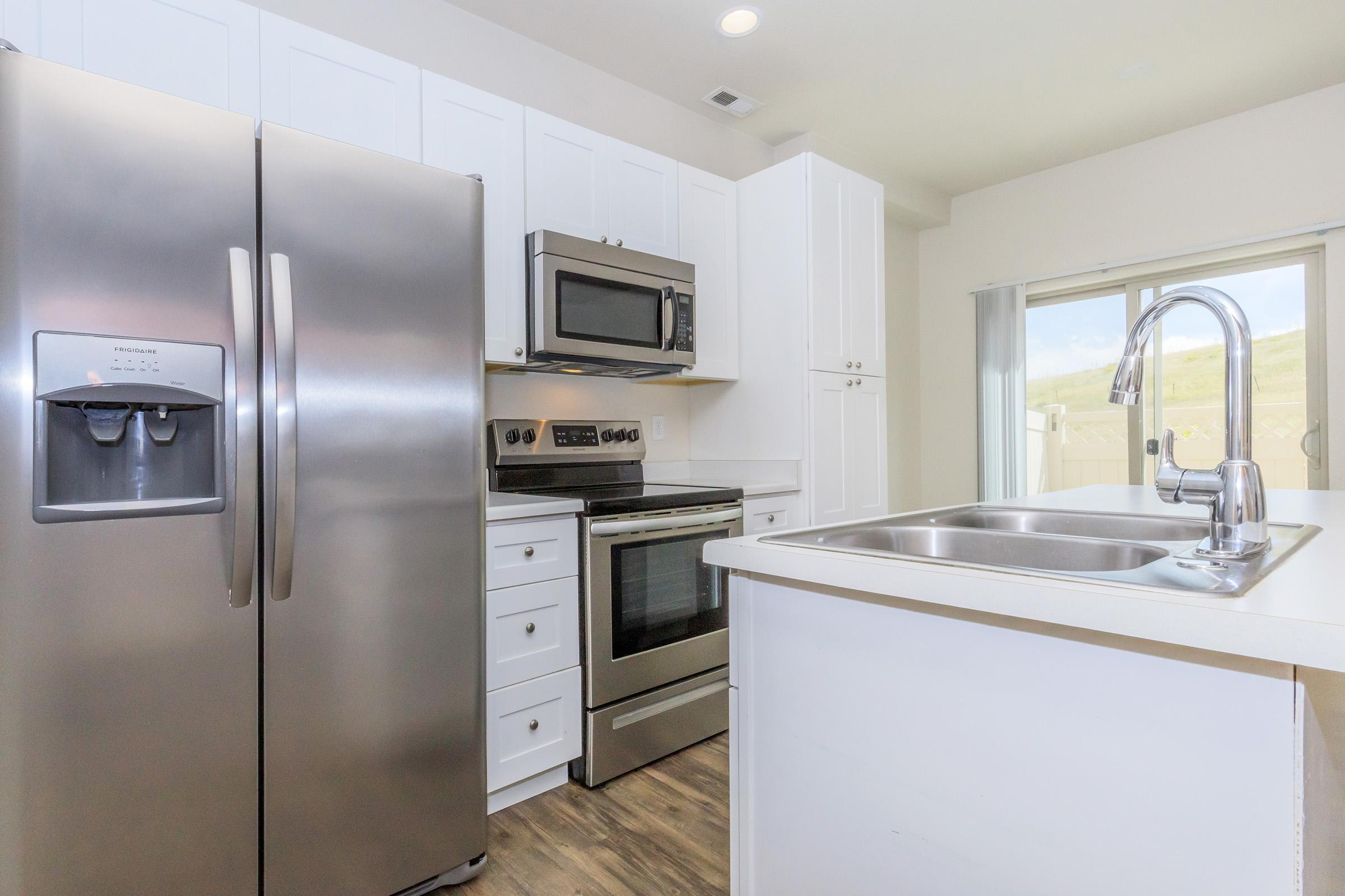 Modern kitchen featuring stainless steel appliances, including a refrigerator, microwave, and oven. The cabinetry is white, complemented by a light countertop and a double sink. Natural light streams in through a nearby window, illuminating a clean and spacious layout.