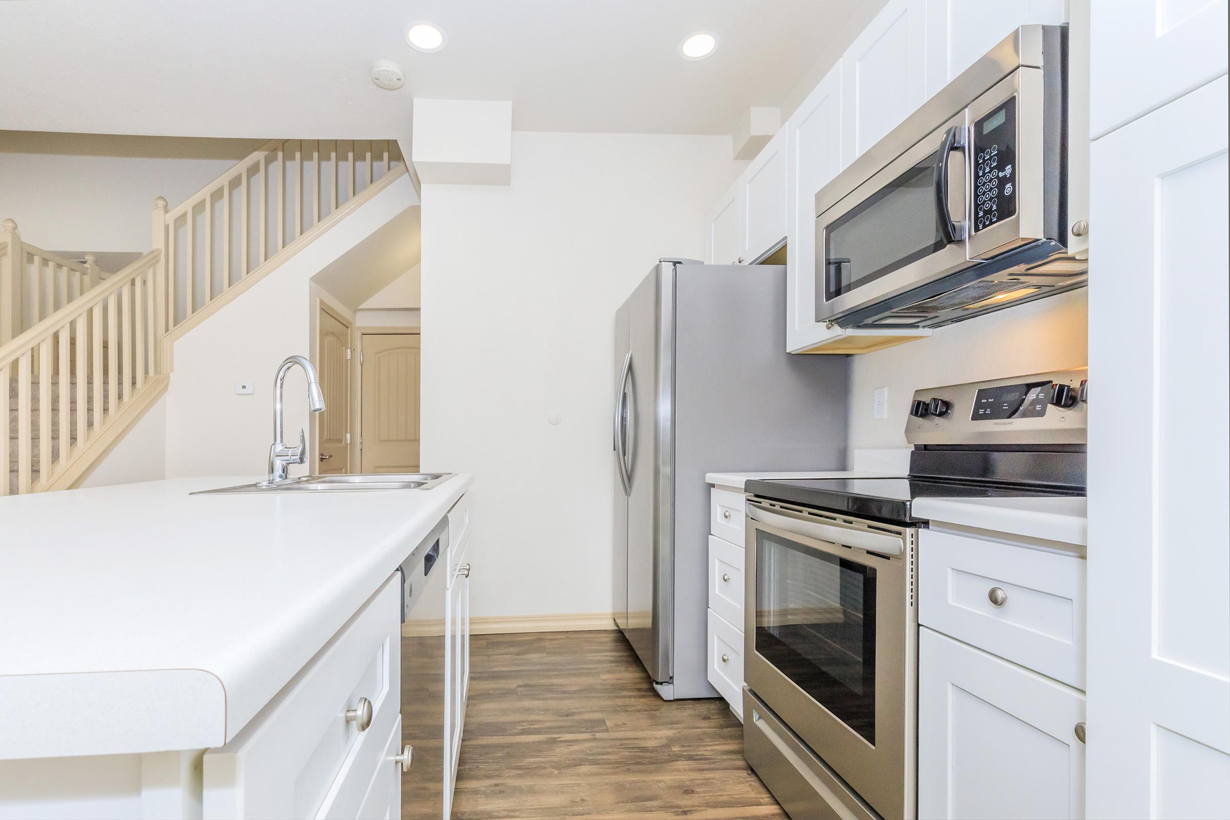 A modern kitchen featuring white cabinetry, stainless steel appliances including a refrigerator, microwave, and oven. There is a large island with a sink, and the space is well-lit with recessed ceiling lights. A staircase is visible in the background, leading to another level.