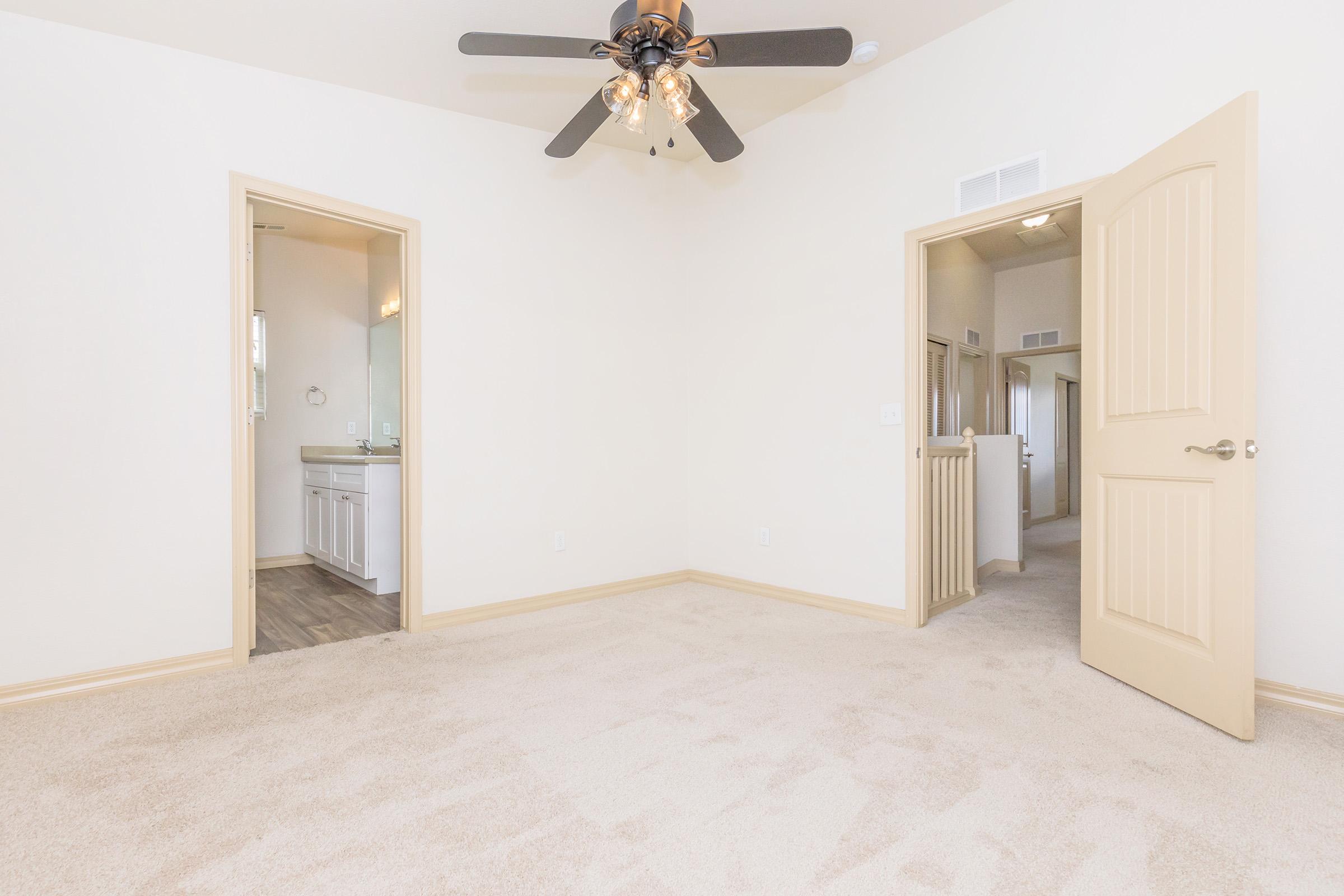 A spacious, empty room with beige carpet and light-colored walls. There is a ceiling fan with lights, two doorways leading to other areas, including a bathroom visible in one doorway. Natural light fills the space, creating a bright and inviting atmosphere.