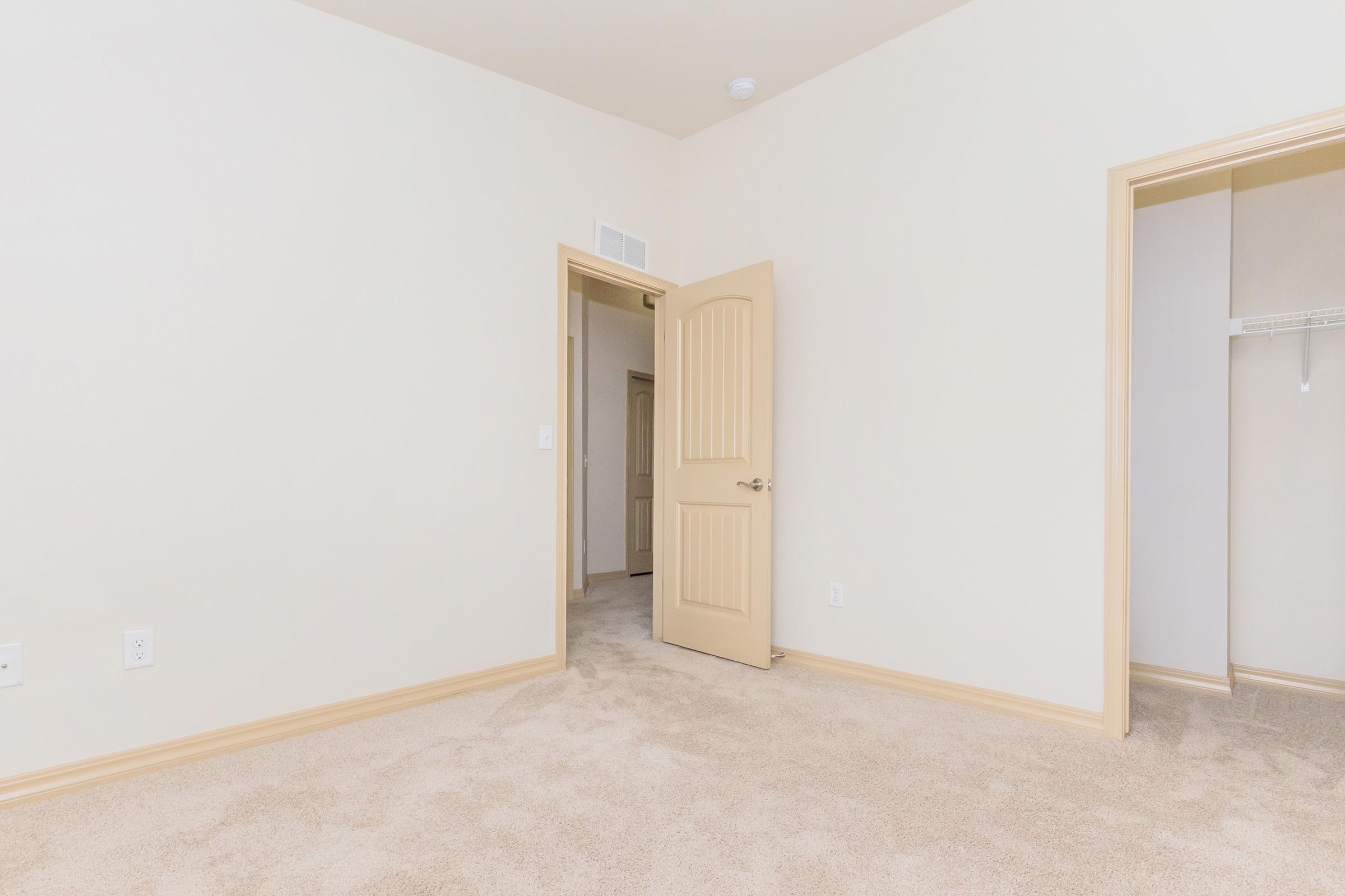 Empty room with light beige walls and carpet, featuring a partially open door leading to a hallway. A closet with a hanging rod is visible on the right side, while natural light illuminates the space, giving a spacious and neutral atmosphere.