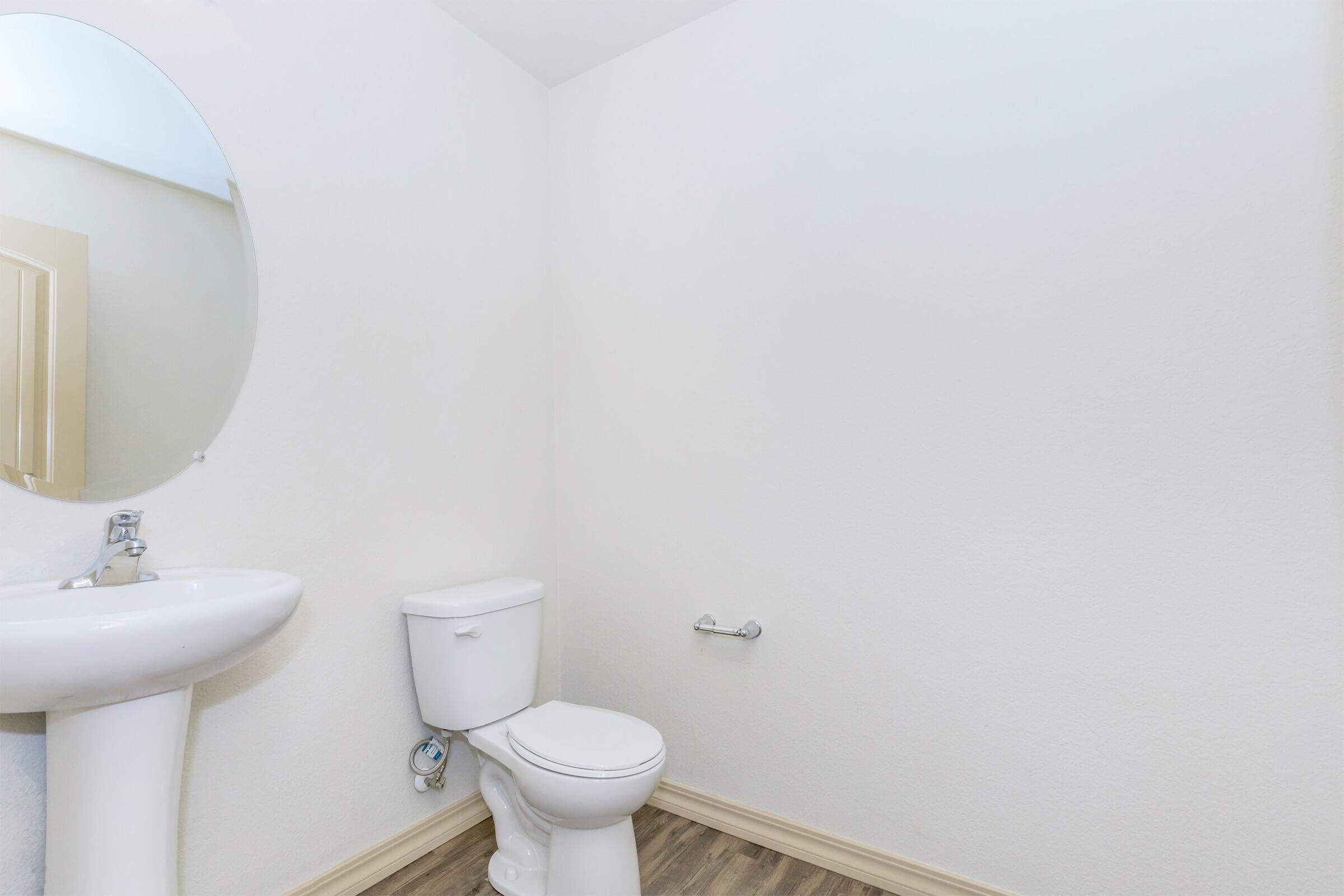 A clean, minimalistic bathroom featuring a round mirror above a white sink, a white toilet, and a wall-mounted toilet paper holder. The walls are painted white, and there is light-colored flooring. The overall atmosphere is bright and airy.
