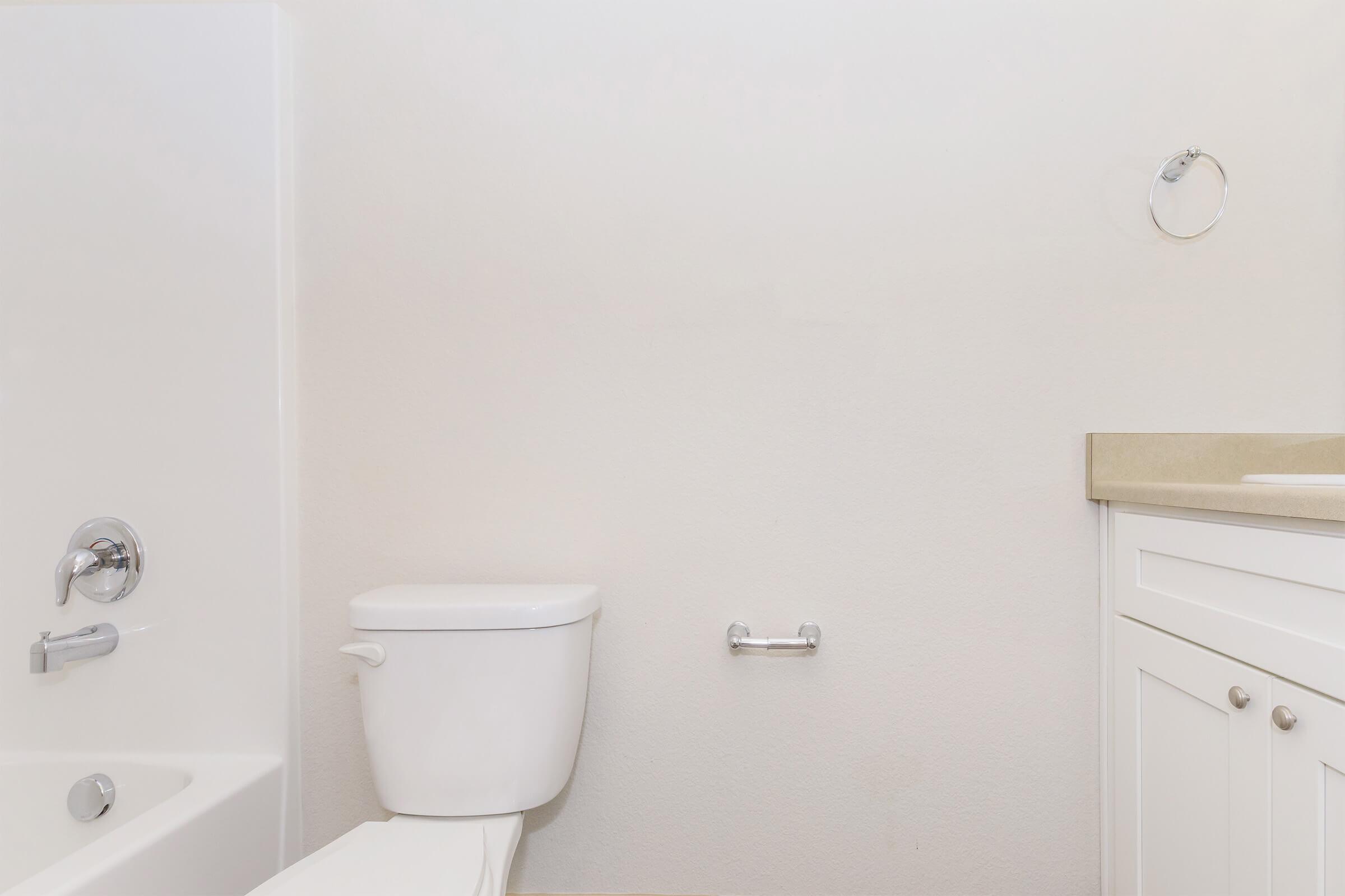A clean, minimalistic bathroom interior featuring a white toilet, a bathtub, and a sink area with a light-colored countertop. The walls are painted in a soft white, and there is a towel rack on the wall. The overall design emphasizes simplicity and modernity.