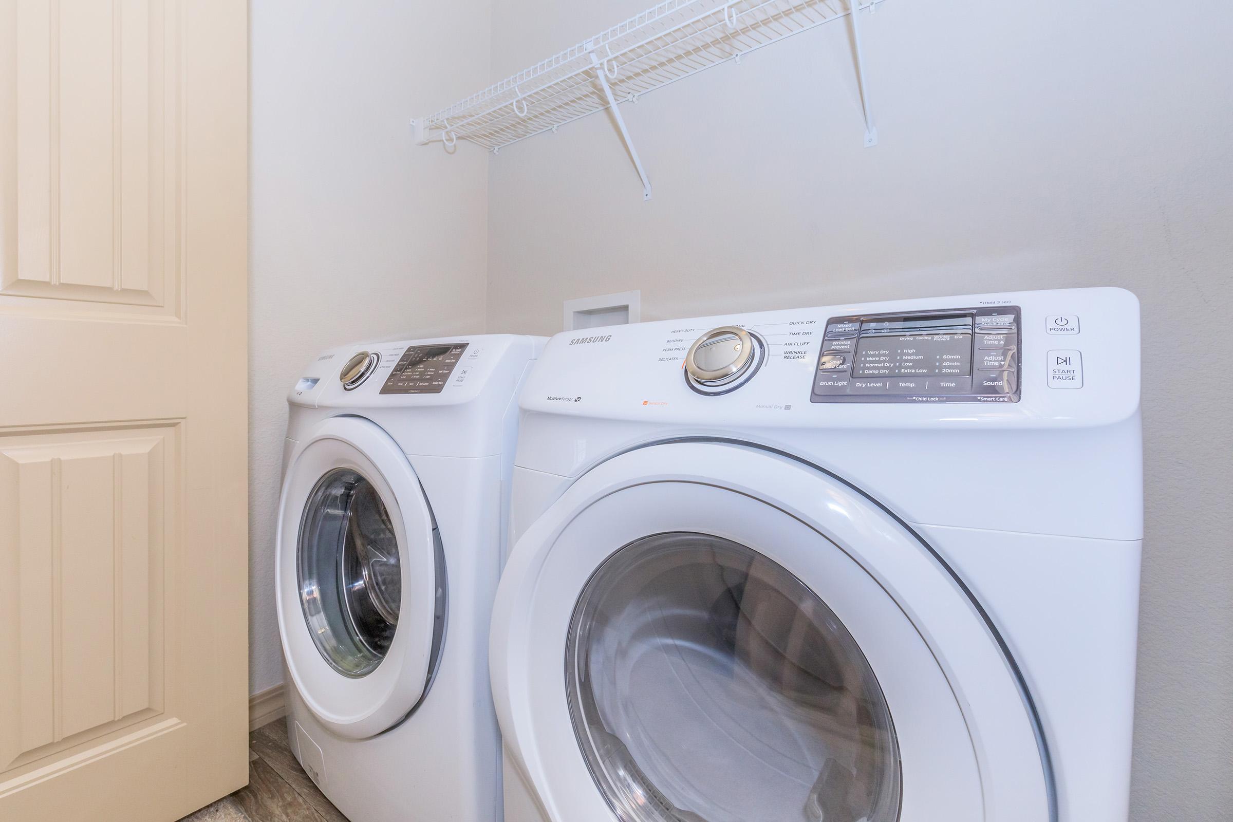 A bright laundry room featuring two white front-loading washing machines side by side. The machines have digital control panels and glass fronts, showcasing their sleek design. Above them, a white wire shelf is mounted on the wall, adding storage space, while a light-colored door is partially visible in the background.