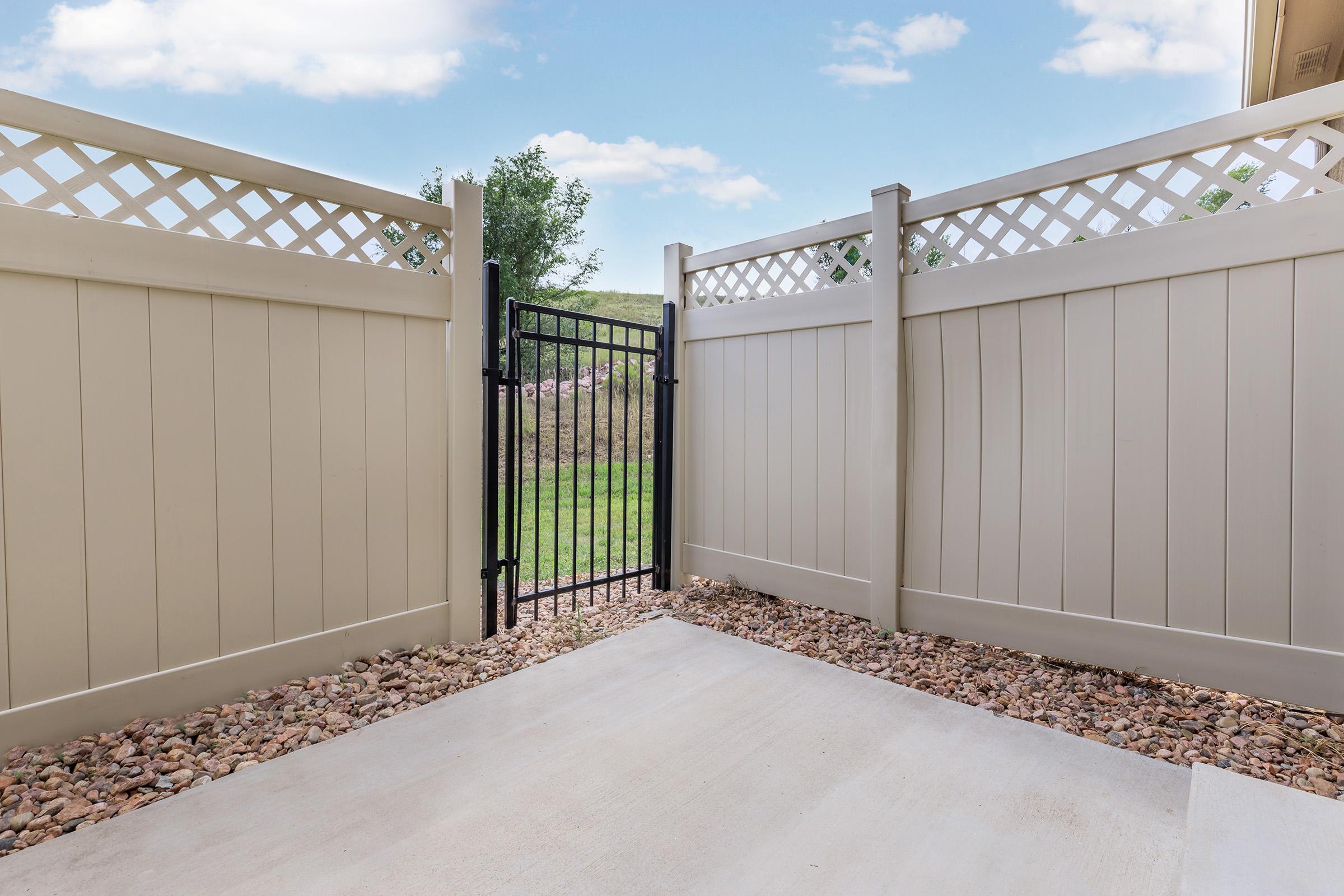 A view through a black metal gate in a fenced area, bordered by beige vinyl fencing with a decorative lattice on top. The ground is covered with small stones, and green grass is visible beyond the gate under a blue sky with a few clouds.
