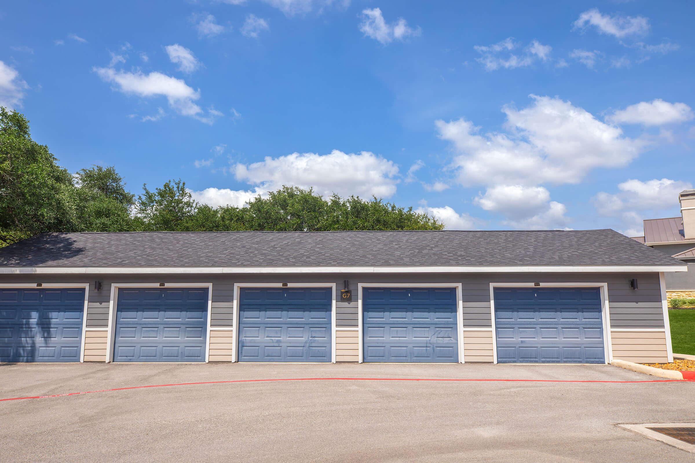 A row of five garage doors with blue panels and gray siding, located in a suburban area. The sky is partly cloudy, with patches of blue visible among the clouds. Green trees are seen in the background, and the driveway is gray with a red edge.