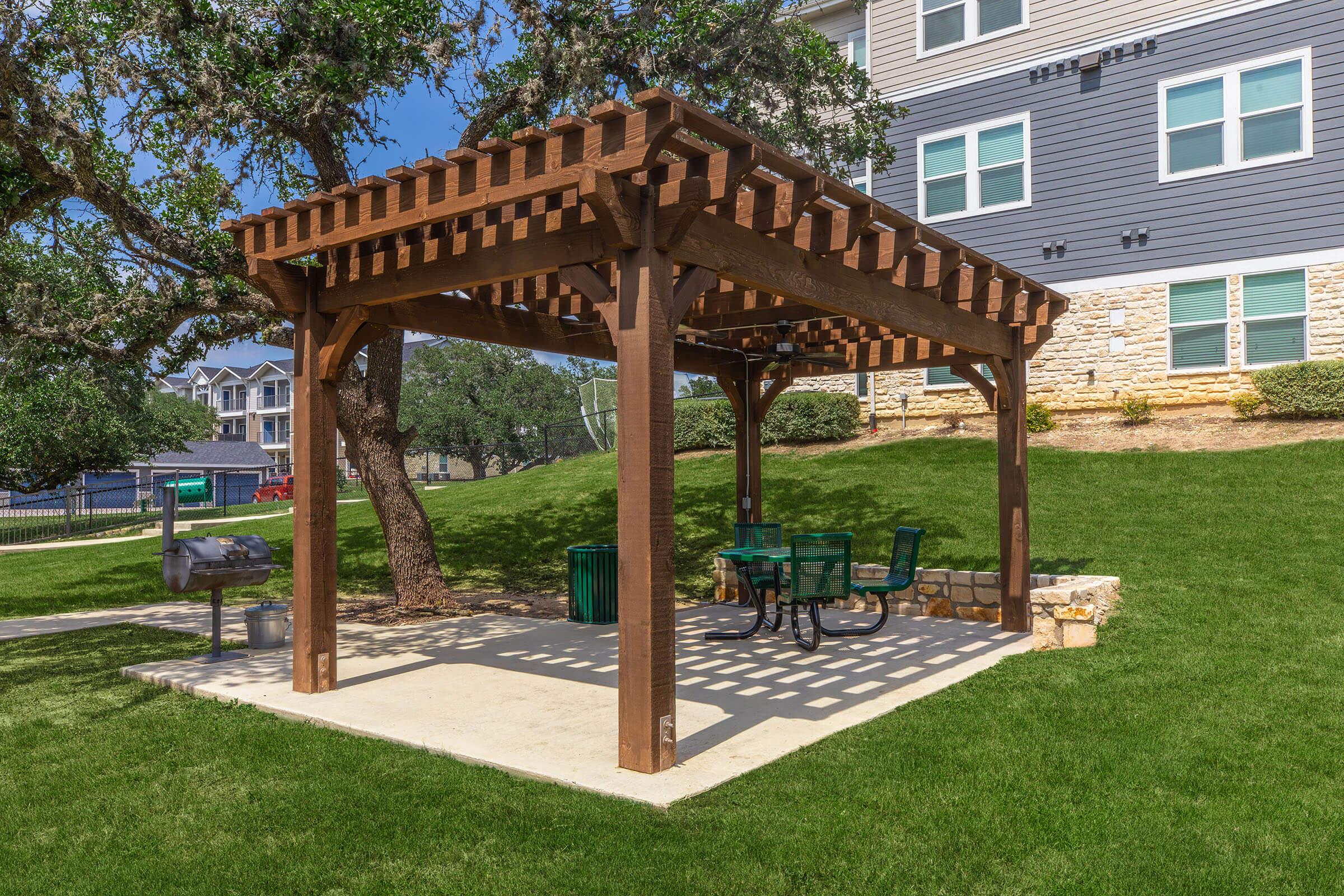 A wooden pergola with a slatted roof provides shade over a picnic area featuring a table and chairs. The structure is set on a grassy area, with nearby trees and an apartment building in the background. A grill and trash can are also visible.