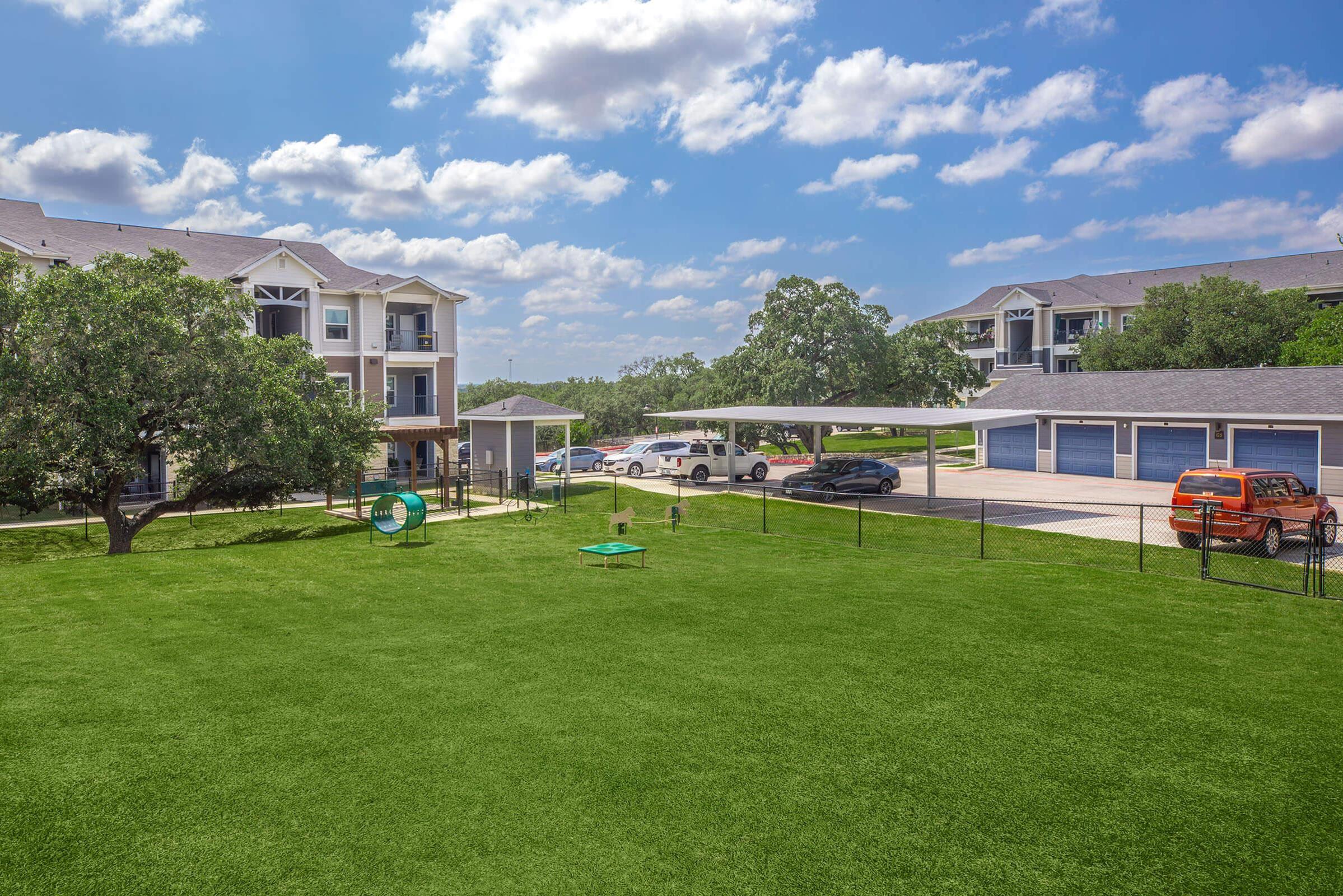 A residential area featuring multiple two-story apartment buildings surrounded by green lawns. In the foreground, there's a playground structure and a fenced area, with a parking lot showing several cars parked underneath covered spaces. The sky is partly cloudy, creating a bright atmosphere.