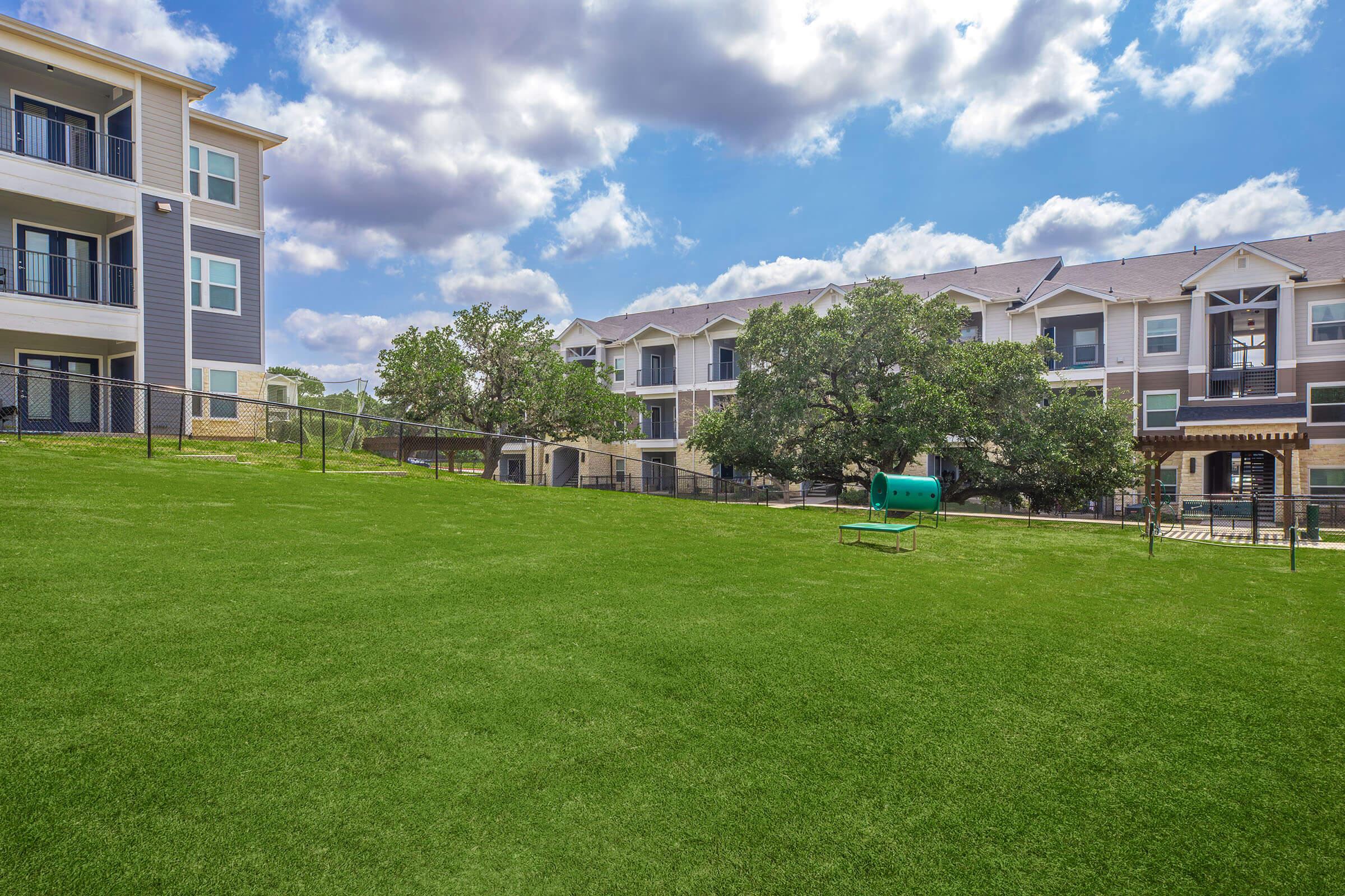 A green grassy area in an apartment complex, featuring two sets of buildings. One building is painted in gray and white, while the other has a similar architectural style. Blue sky with fluffy clouds overhead and a small dog station in the foreground.