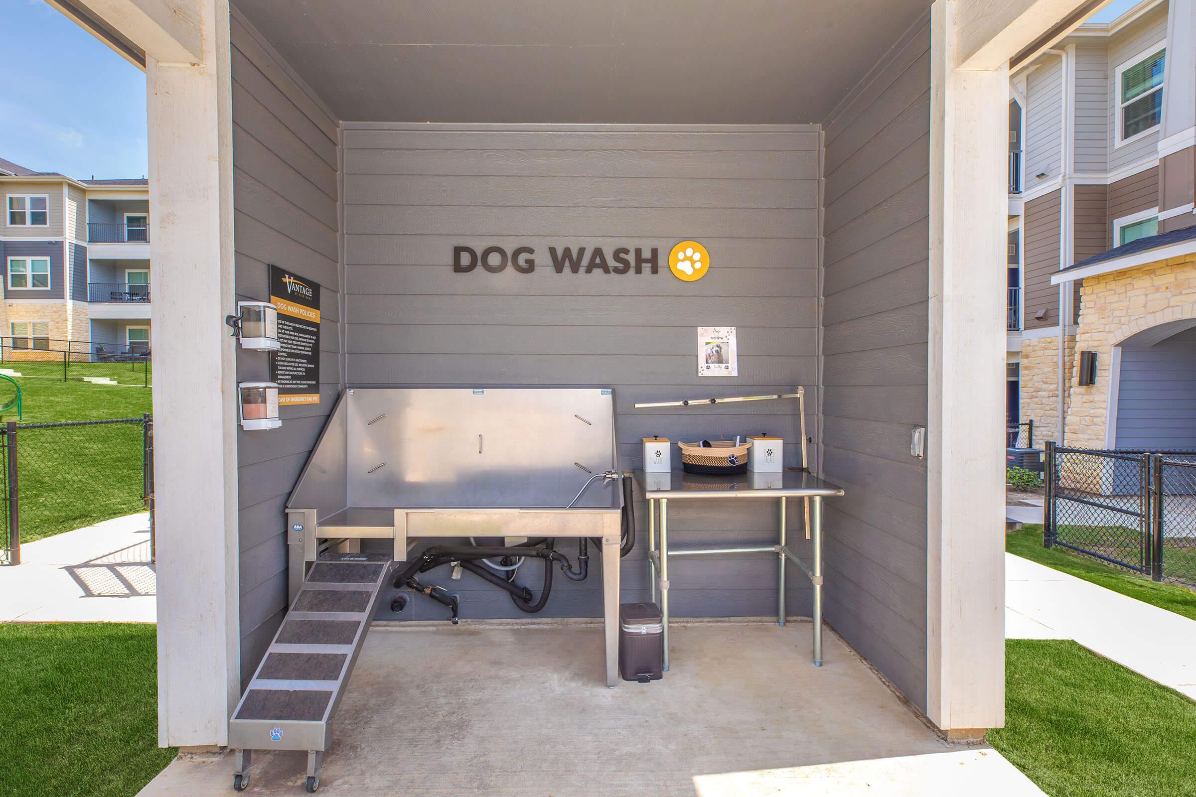 A clean and well-maintained dog wash station enclosed by gray walls. It features a stainless steel washing area with steps, a grooming table, and pet supplies. The words "DOG WASH" are prominently displayed on the wall, alongside a bulletin board. The area is surrounded by green grass and nearby buildings.