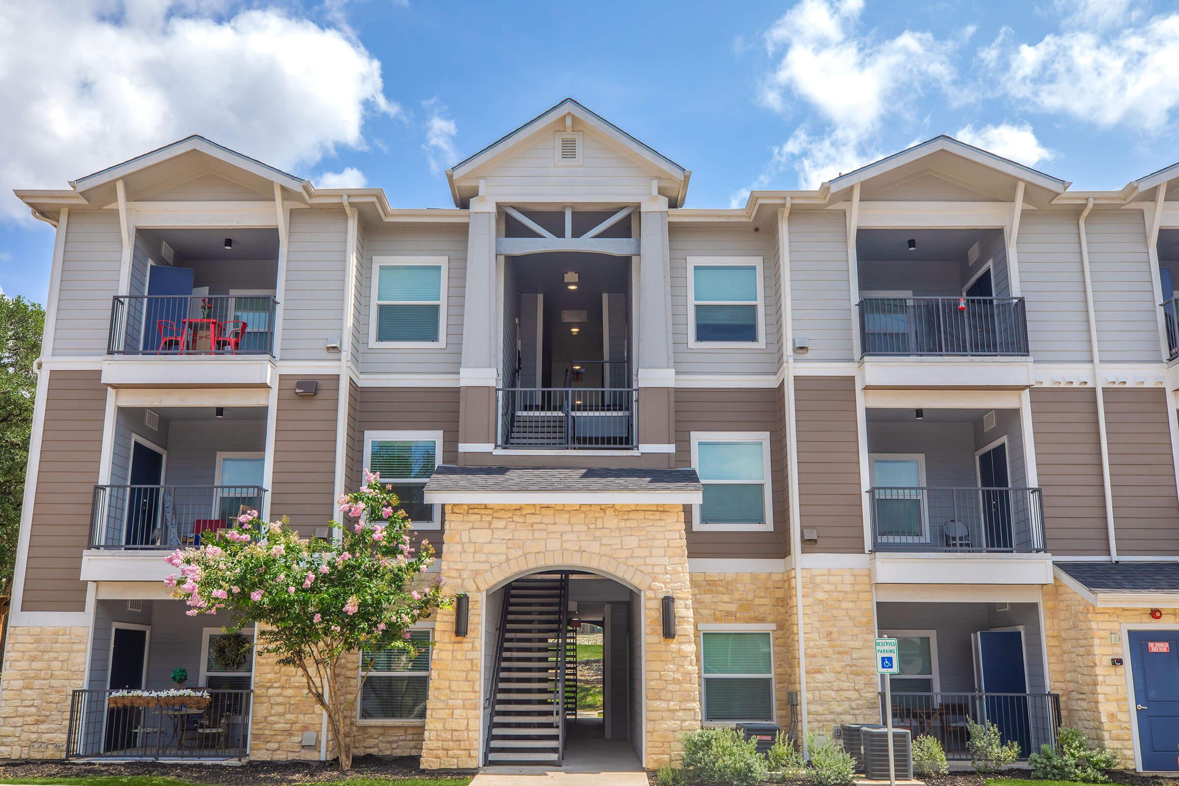 A three-story apartment building with a stone base and light-colored siding. The entrance features a set of stairs leading to balconies with seating. Lush greenery and blooming flowers are in the foreground, with blue skies and fluffy clouds above. Balconies are adorned with patio furniture.