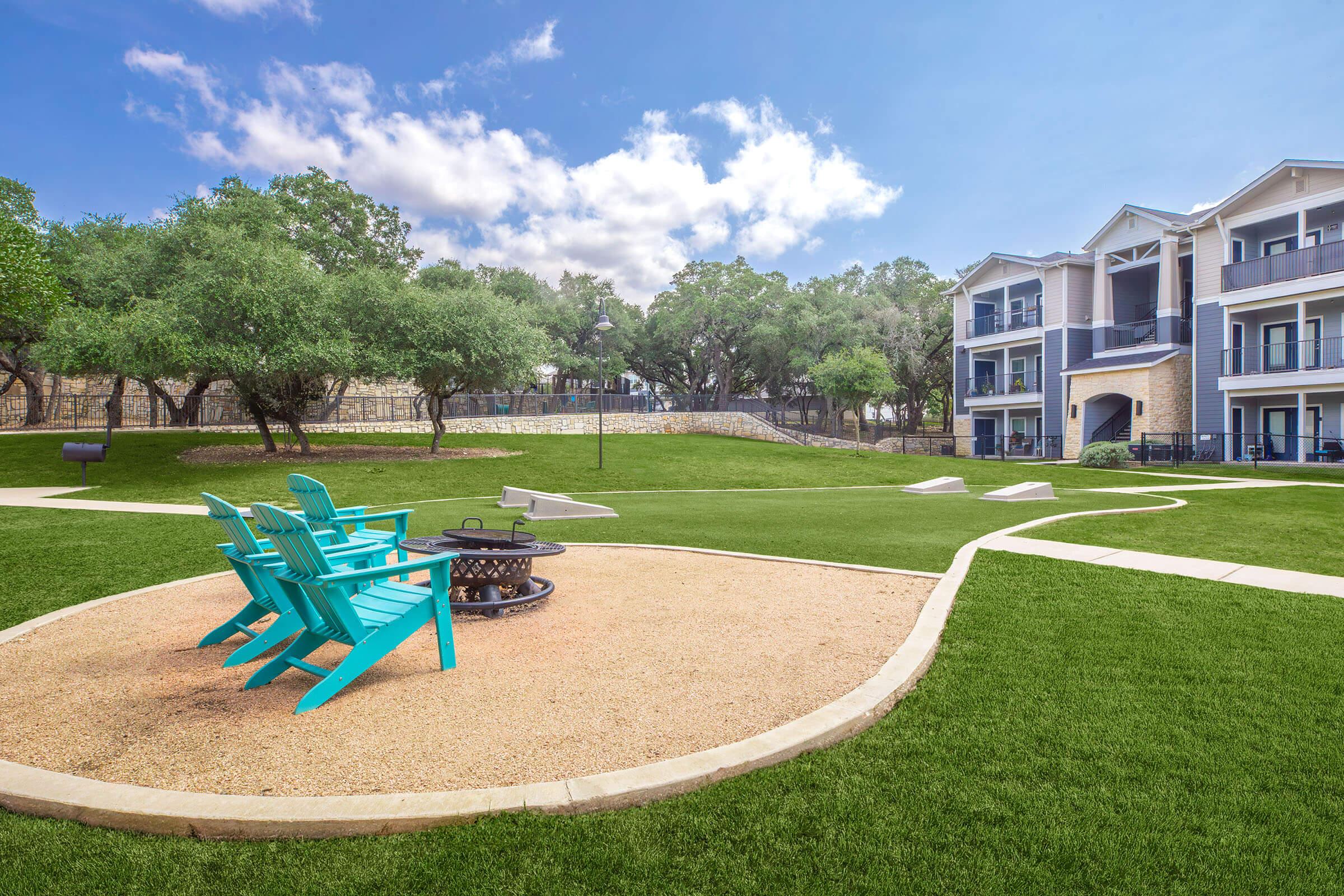 A peaceful outdoor area featuring vibrant teal Adirondack chairs arranged around a fire pit, set on a manicured lawn. In the background, there are modern apartment buildings and trees with a clear blue sky overhead, creating a serene and inviting atmosphere for relaxation.