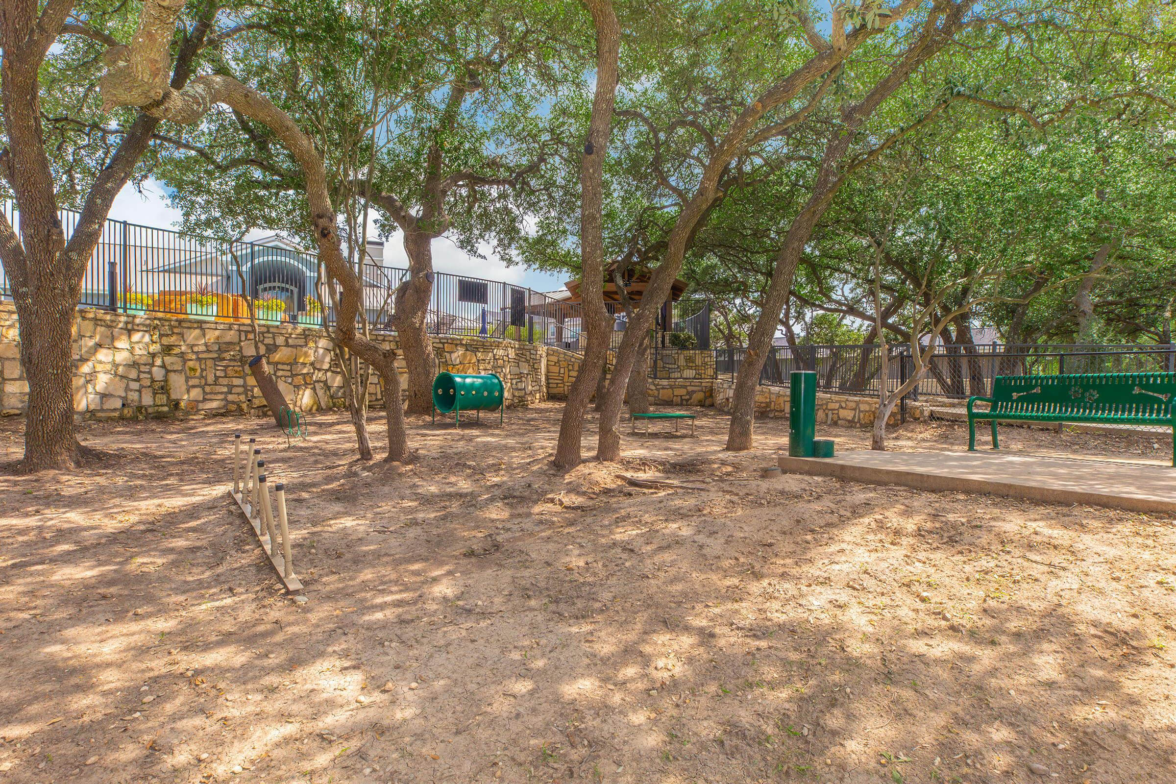 A park scene featuring a shaded area with several trees, a small playground structure in the background, a green bench, and a playground climbing structure. The ground is sandy and there is a low wooden obstacle for kids to play on. The atmosphere is sunny and inviting, perfect for outdoor activities.