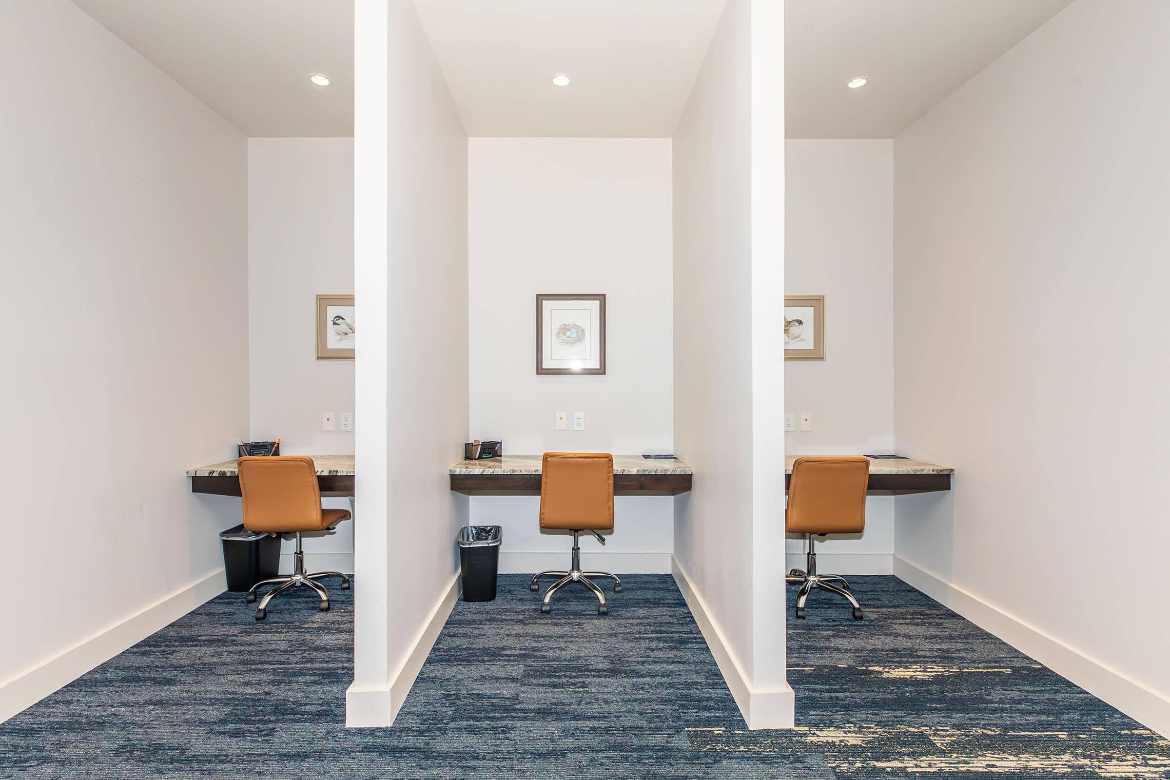 Two separate workstations with modern chairs in a minimalist office environment. Each station features a small desk, lamp, and decorative wall art. The floor is covered with blue carpet, and the walls are painted white, creating a clean and open atmosphere.
