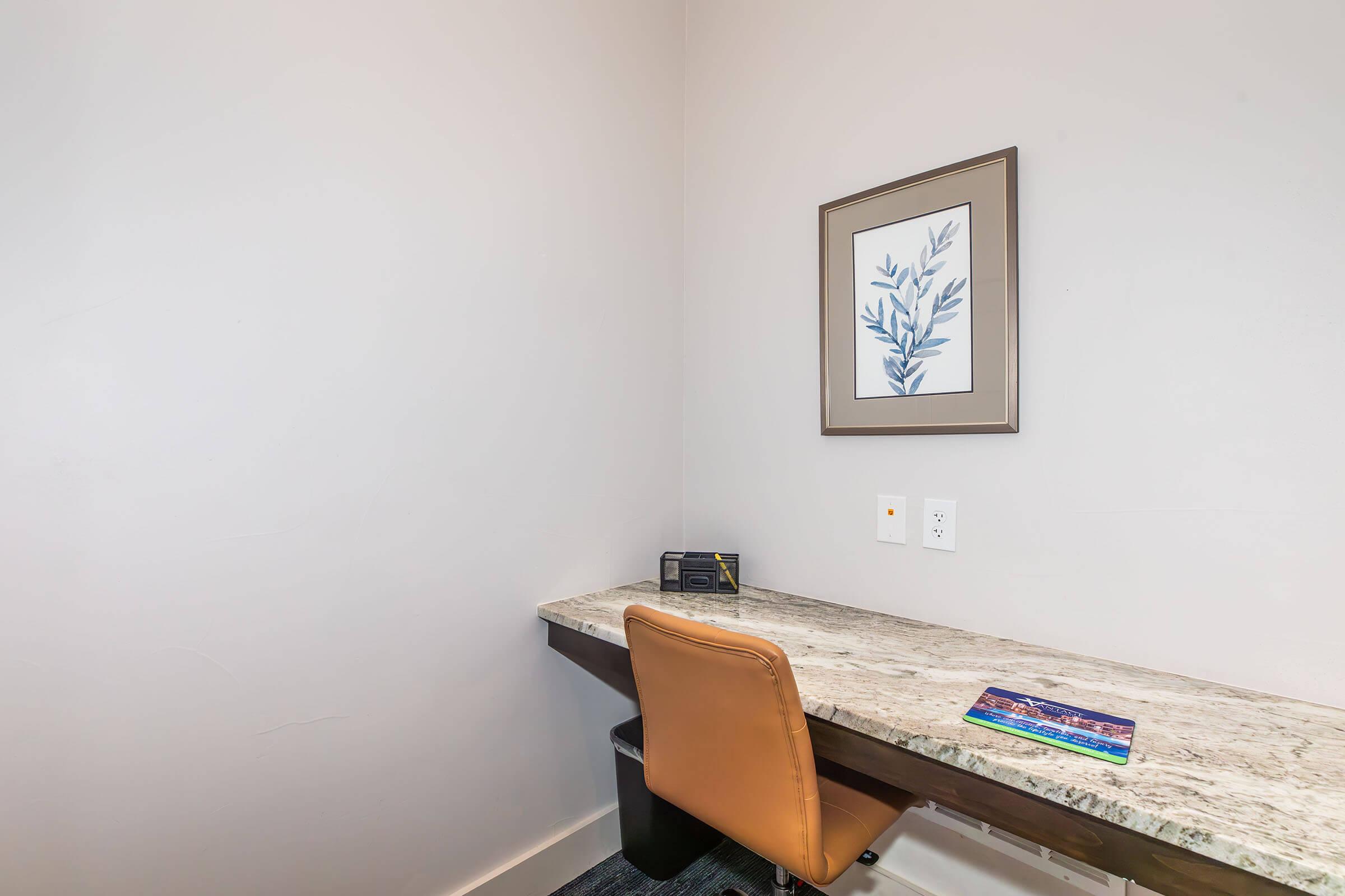 A small workspace in a room featuring a granite-topped desk, an orange chair, a black trash can, a framed botanical print on the wall, and a magazine on the desk surface. The wall is painted a light color, creating a minimalistic and modern atmosphere.