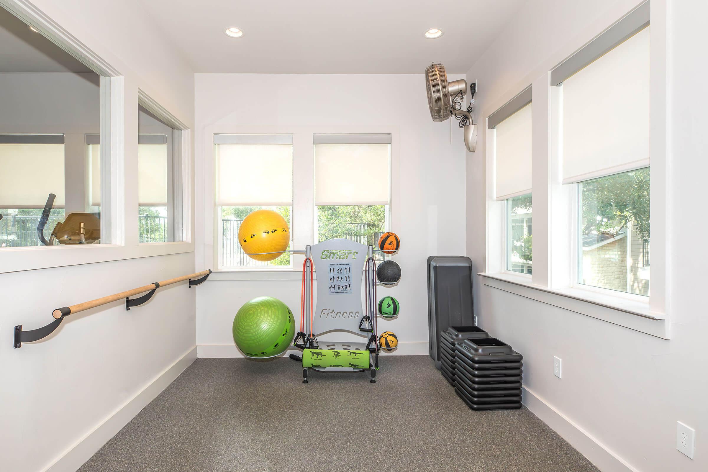 A bright fitness room featuring a wooden barre for stretching, exercise equipment including stability balls and a step platform, along with stacked black risers. Large windows provide natural light, and overhead lights illuminate the space.