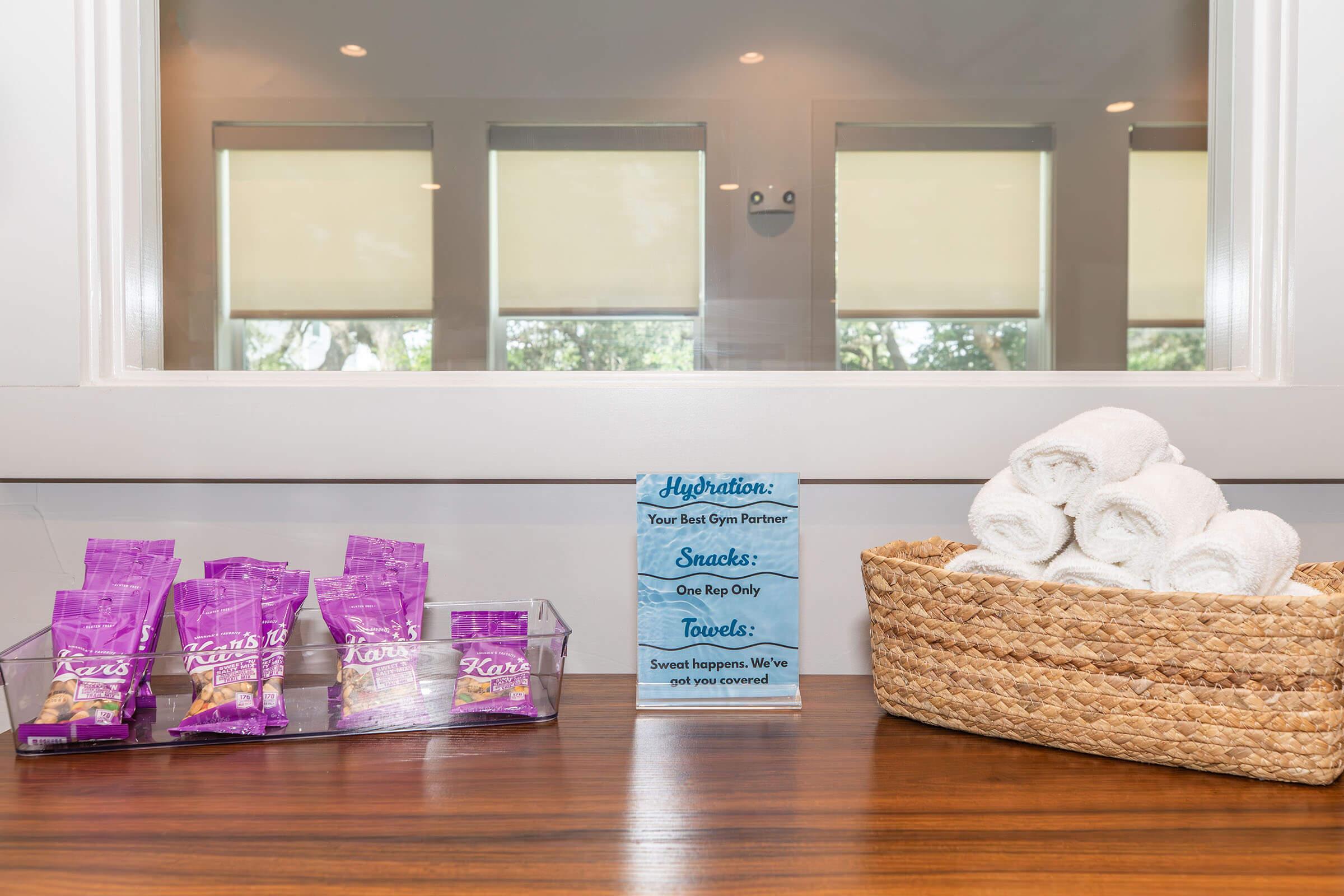 A countertop featuring a basket of neatly rolled white towels, a sign that reads "Hydration: Your Best Gym Partner" alongside snack packs labeled "Kara's" in purple packaging. The background includes large windows with soft shades, creating a bright and inviting atmosphere.