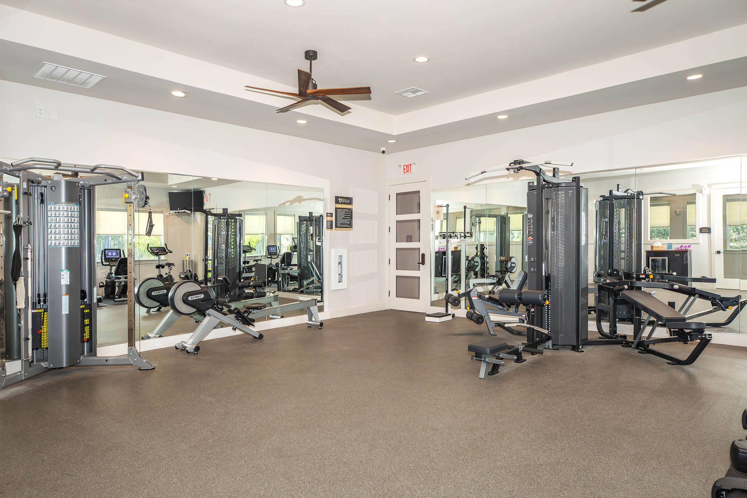 A modern gym interior featuring various exercise equipment, including weight machines, a rowing machine, and a multi-functional workout station. Large mirrors line the walls, and the space is well-lit with ceiling lights and a ceiling fan, creating an inviting workout environment.