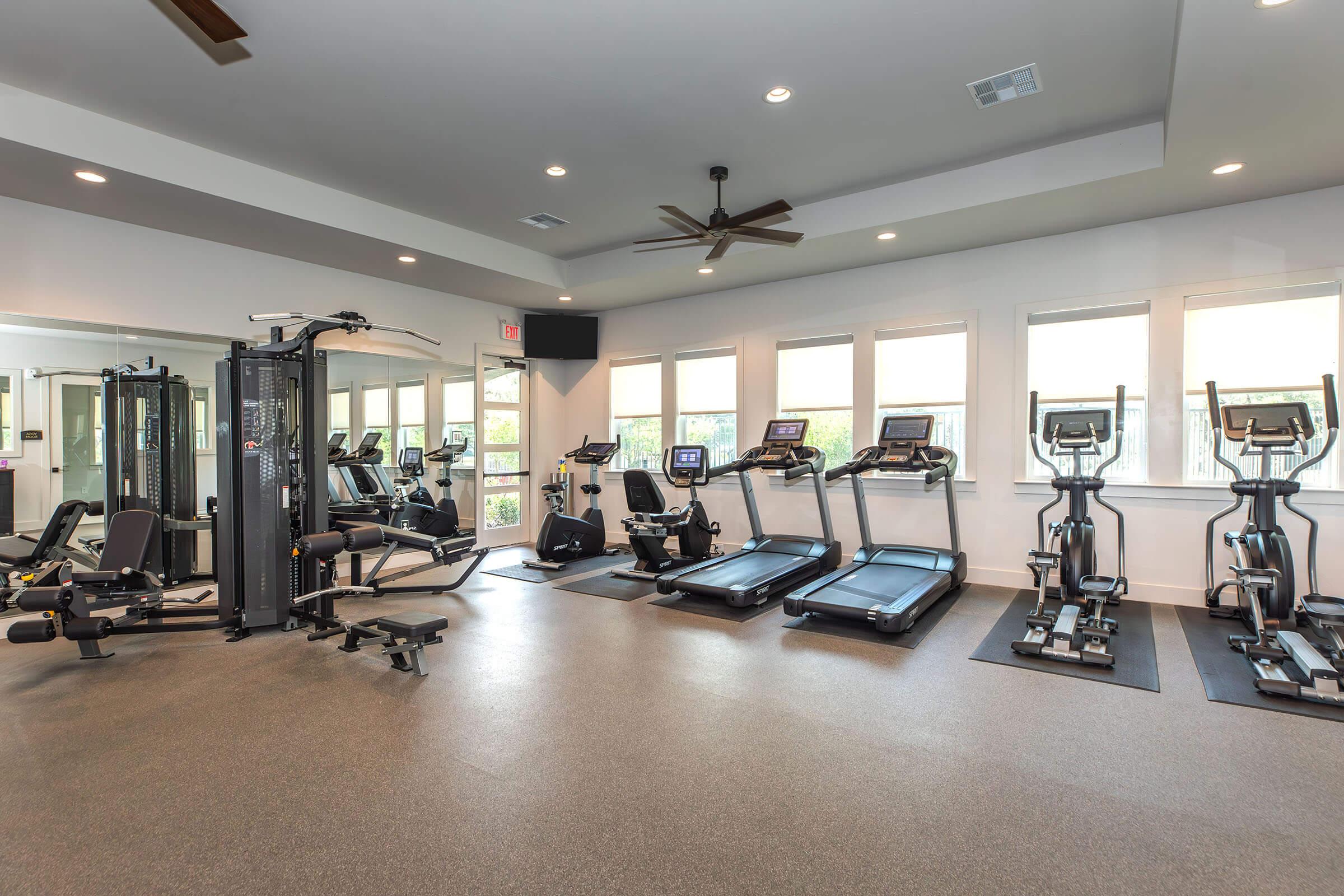 A modern gym interior featuring various exercise equipment including treadmills, elliptical machines, weight machines, and free weights. Large windows provide natural light, and there are ceiling fans for ventilation. The flooring is a dark rubber material, creating a functional workout space.