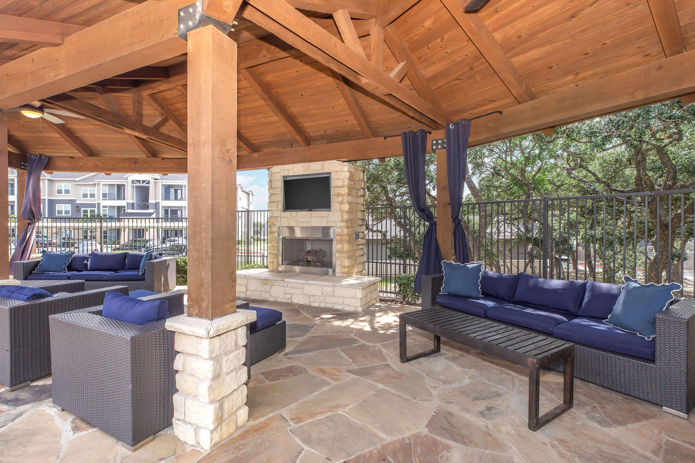 Outdoor seating area featuring a wooden gazebo with a stone fireplace, plush seating in navy blue cushions, and a flat-screen TV mounted on the wall. The space is surrounded by a decorative fence and trees, providing a cozy atmosphere for relaxation.