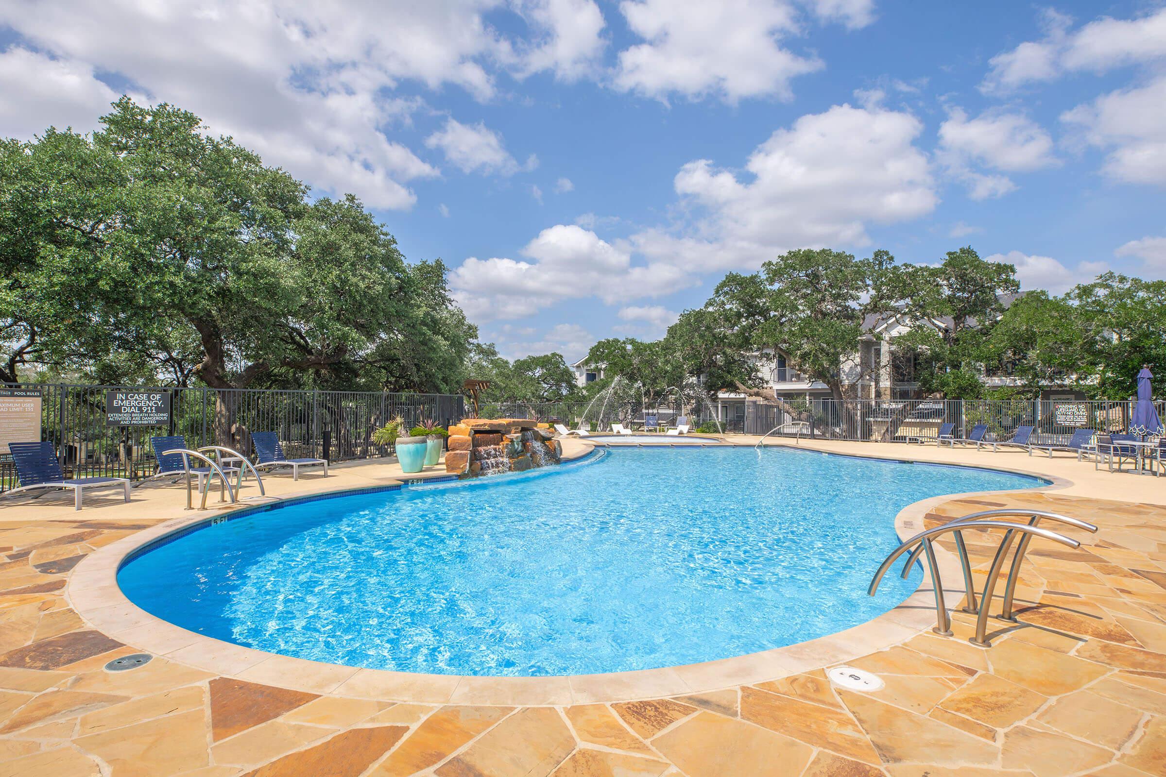 A spacious outdoor swimming pool with clear blue water, surrounded by a stone deck. Lush green trees and a clear blue sky with scattered clouds are visible in the background. The area features lounge chairs and decorative elements, creating a relaxing atmosphere for leisure and enjoyment.