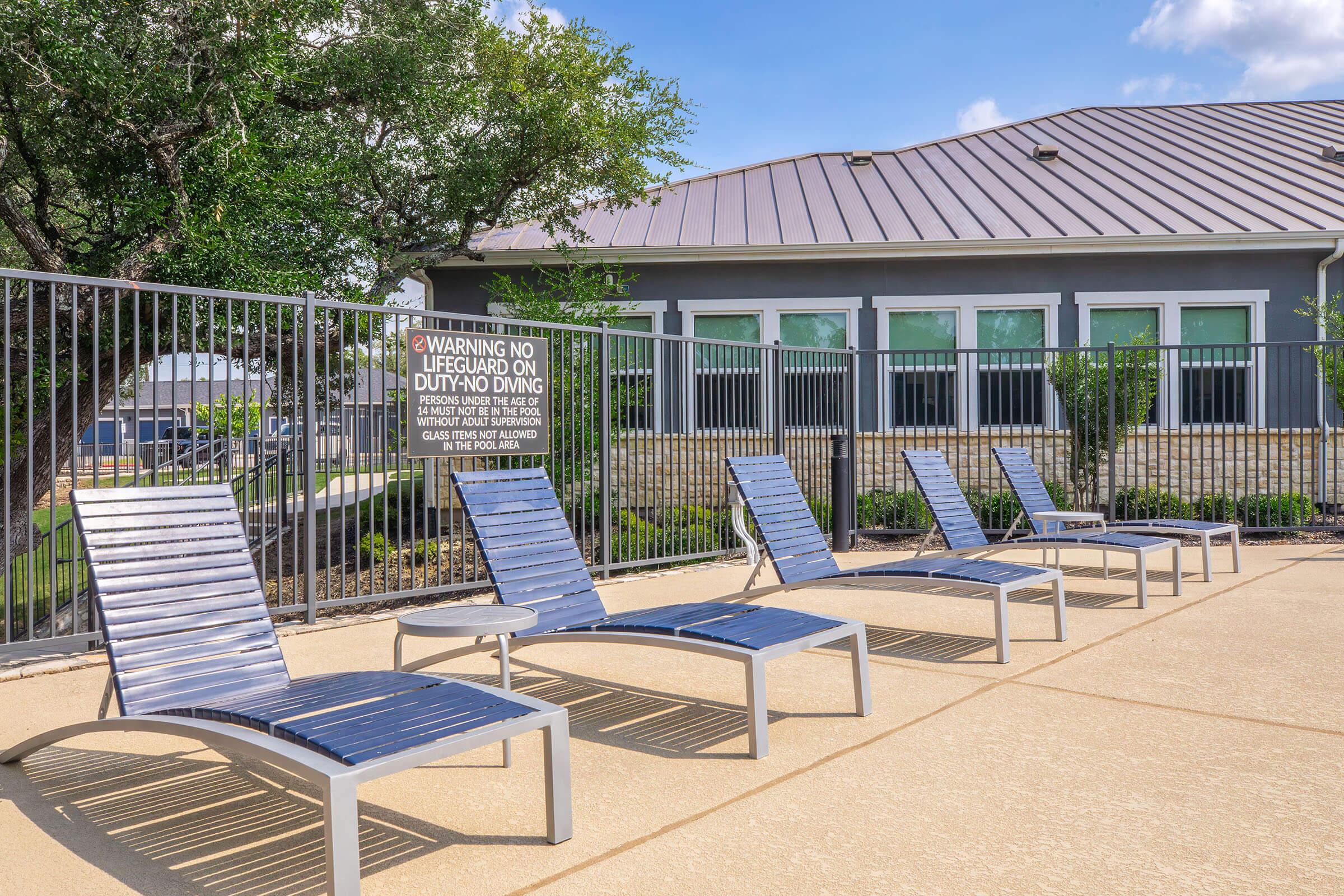 A row of modern, silver lounge chairs by a pool area. The chairs are arranged neatly next to a light-colored concrete deck. In the background, there’s a building with a gray exterior and large windows, along with a safety warning sign regarding lifeguard duties. Green shrubs are visible nearby.