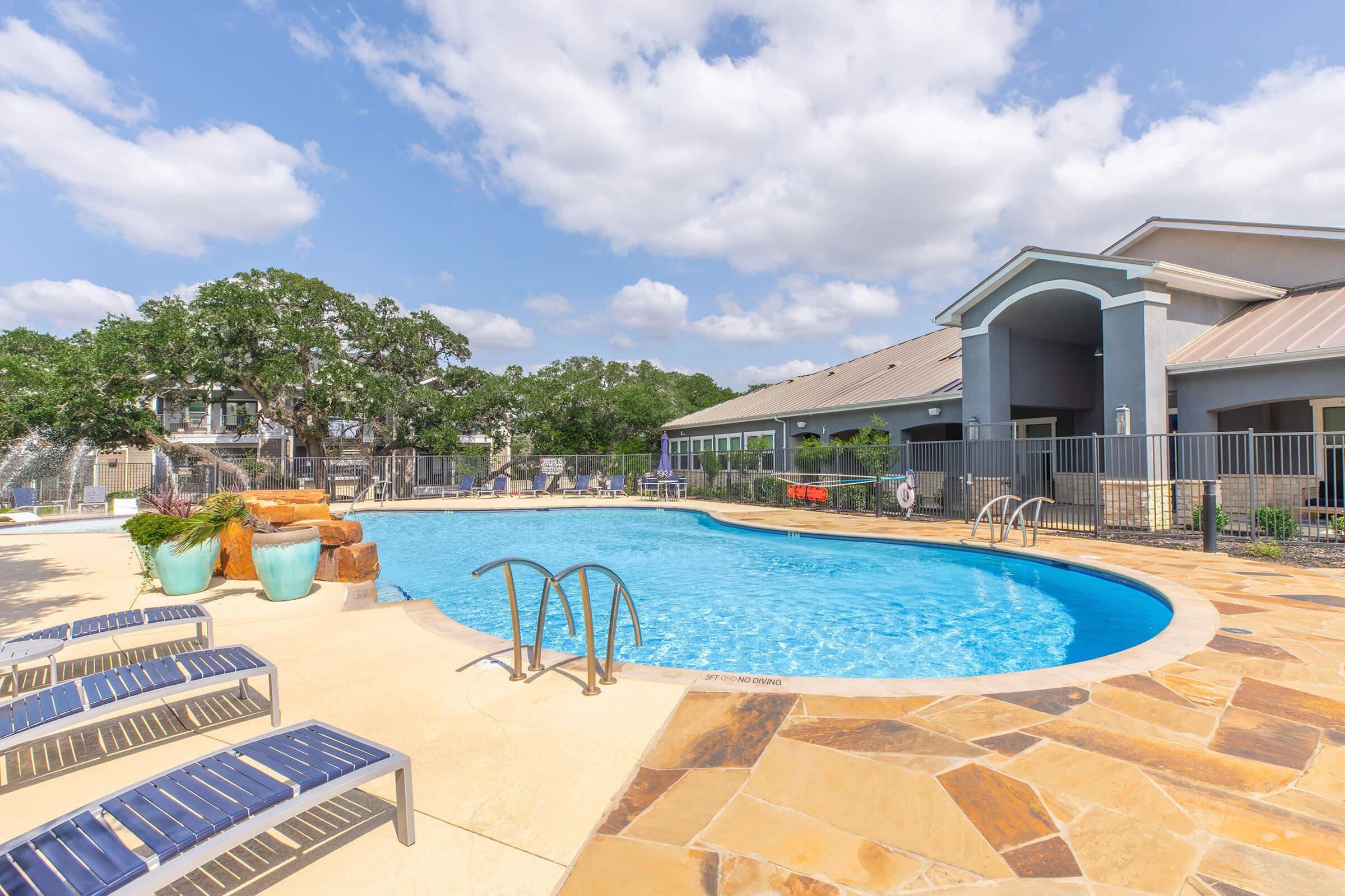 A serene swimming pool surrounded by lounge chairs and landscaped greenery. The pool features a rock waterfall and is bordered by a modern building under a clear blue sky with fluffy clouds. The area has a relaxed, inviting atmosphere, perfect for leisure and relaxation.