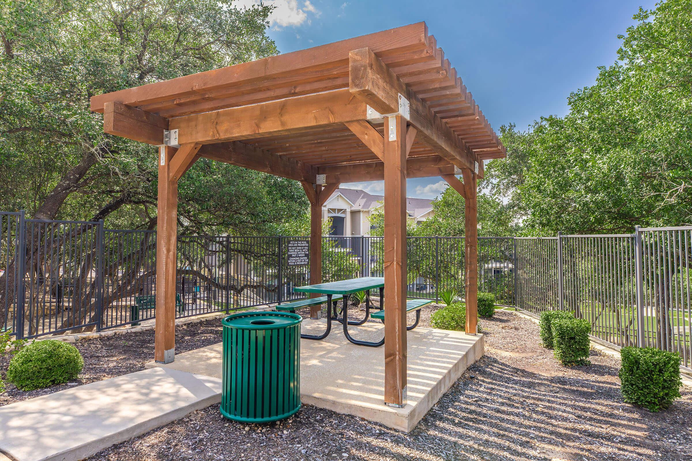 A wooden gazebo with a slatted roof provides shade over a picnic table and green trash can. The structure is surrounded by neatly trimmed shrubs and trees, with a gated area visible in the background and a blue sky above, creating a serene outdoor space.