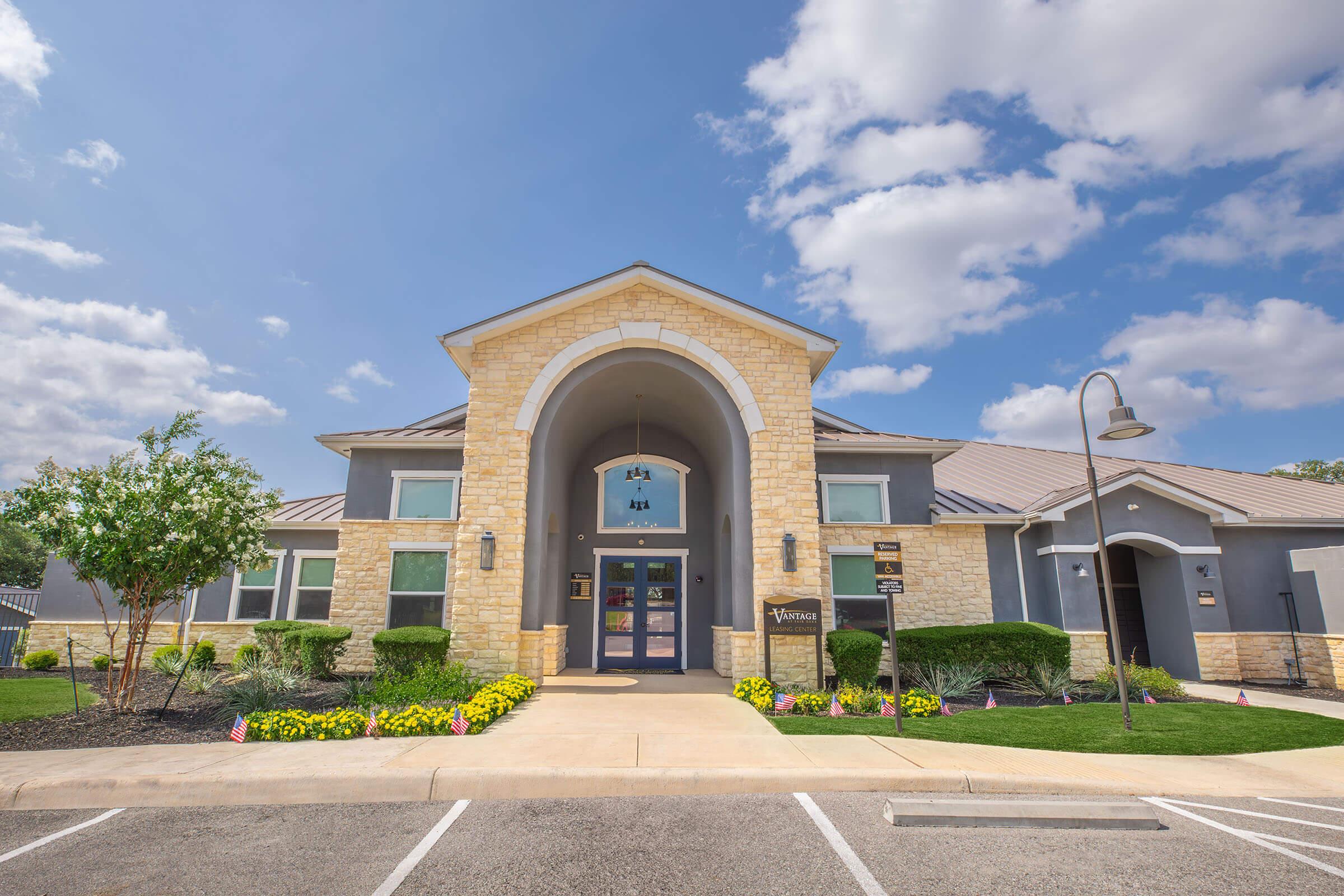A modern building with a stone facade and a large welcoming entrance under a curved arch. The surrounding landscape features green shrubs, colorful flower beds, and manicured lawns, under a partly cloudy blue sky. Parking spaces are visible in the foreground.