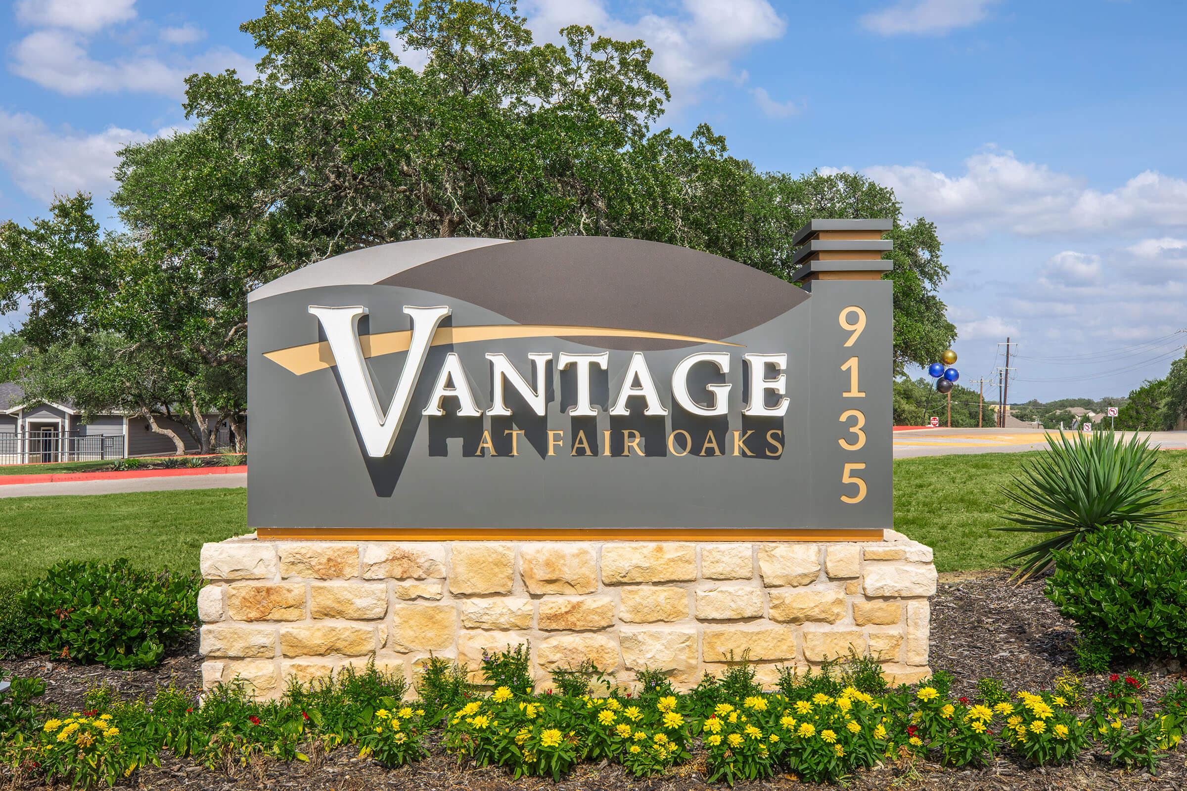 Sign displaying "Vantage at Fair Oaks" with the address "9135," surrounded by landscaped greenery and colorful flowers under a blue sky.