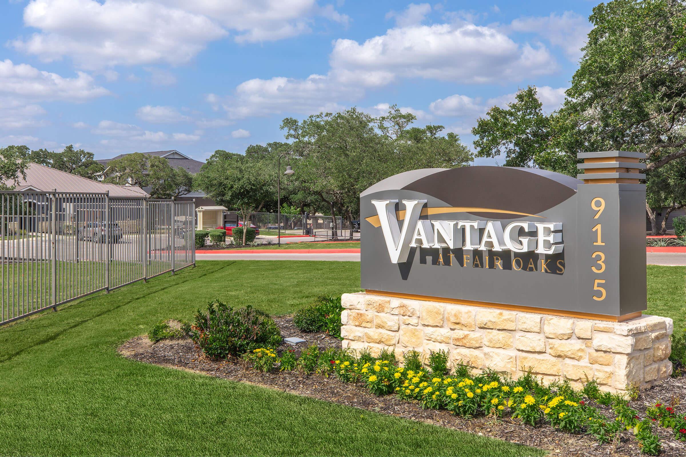 Sign for Vantage at Fair Oaks with the address 9135, surrounded by well-maintained landscaping and a fence in the background. The setting features green grass and decorative flowers, under a partly cloudy sky.